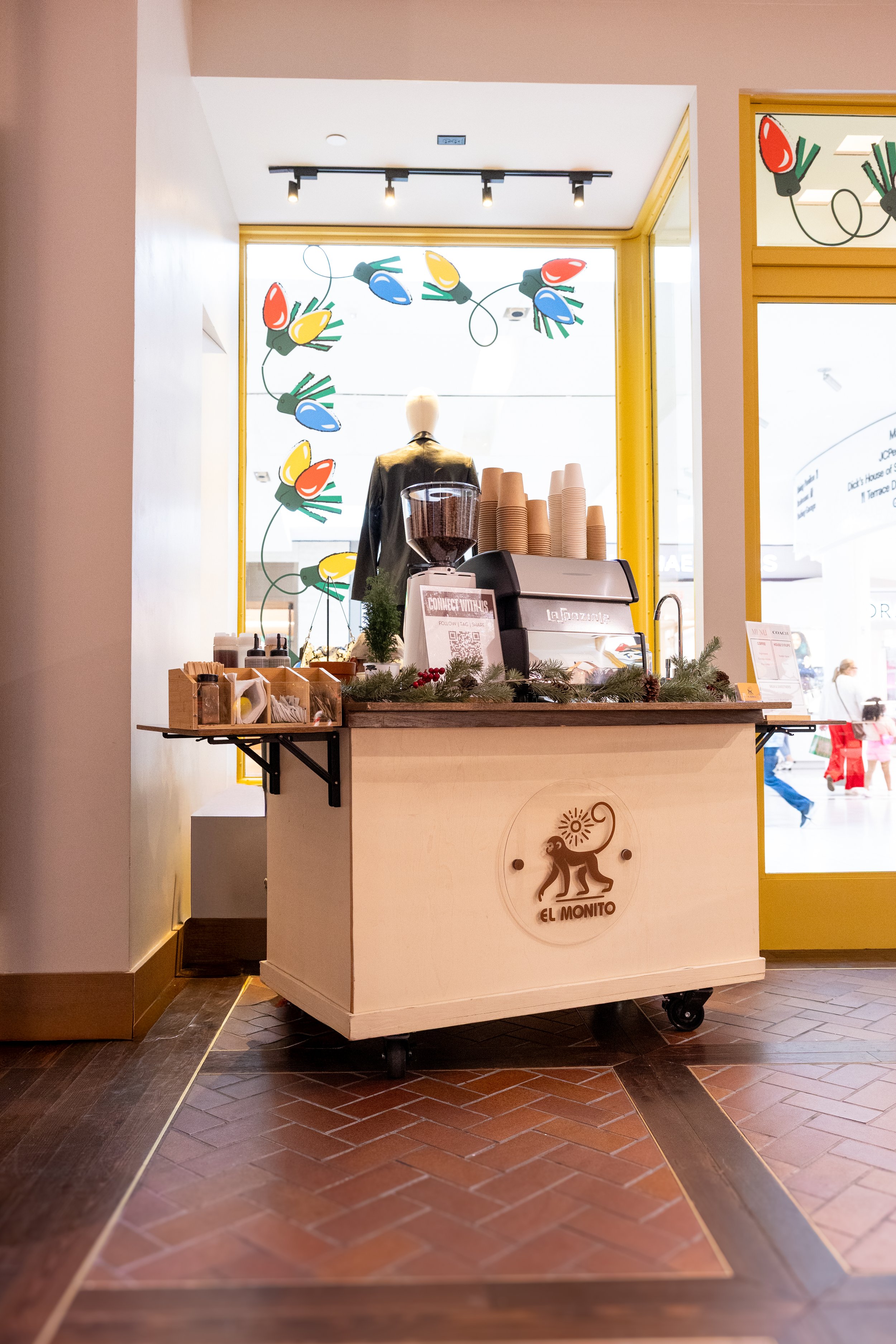 A small mobile coffee cart branded 'El Monito' with a wooden finish, decorated with greenery and berries, positioned near a large window with colorful holiday string lights and a mannequin wearing a jacket in the background.