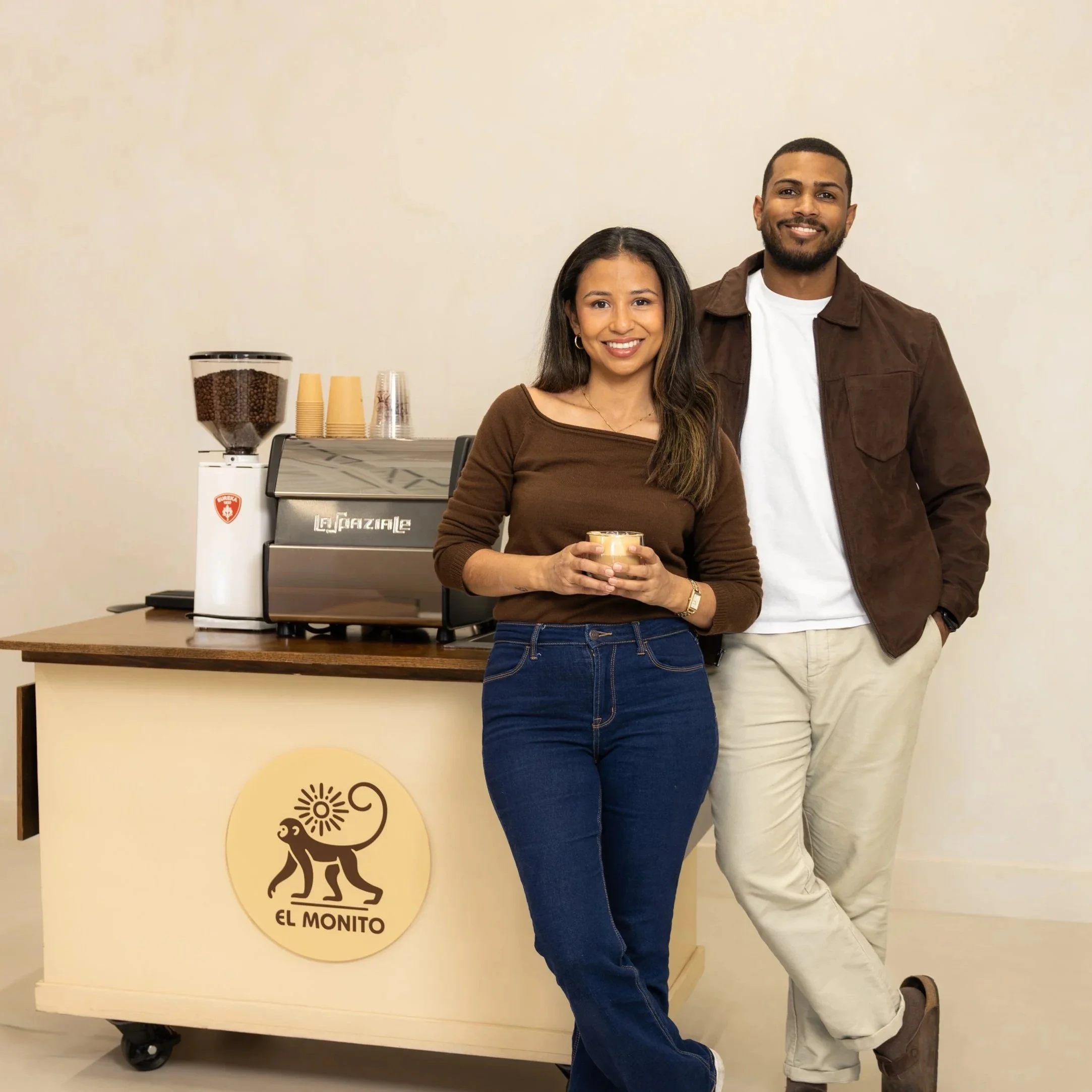 A woman and a man standing in front of a coffee station at El Monito. The woman is holding a coffee cup and smiling, the man has his hands in his pockets and is also smiling.
