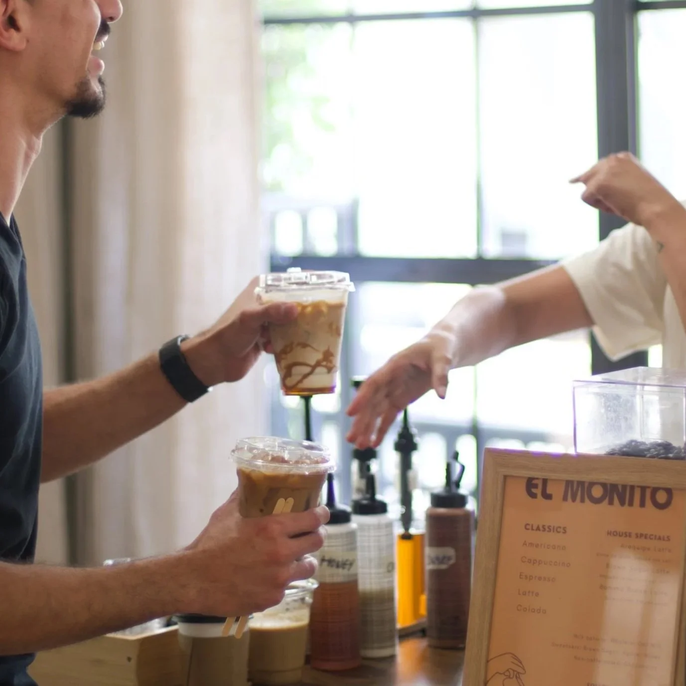 A person handing a coffee drink to another person at a coffee shop, with a menu visible on a stand in the foreground.