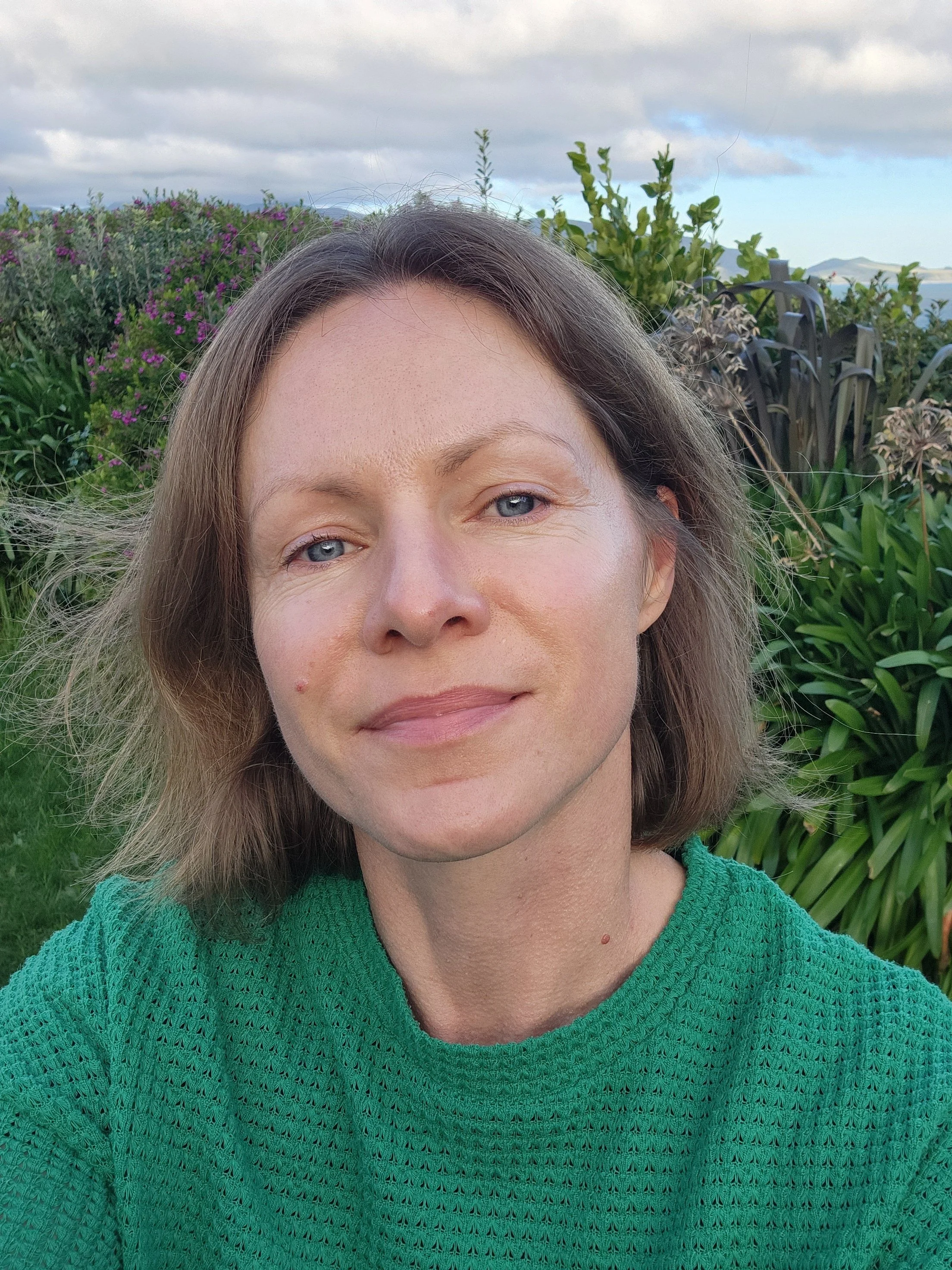 A smiling and confident woman with shoulder-length brown hair and blue eyes taking a selfie outdoors in front of green plants and purple flowers, with a cloudy sky and distant landscape in the background.
