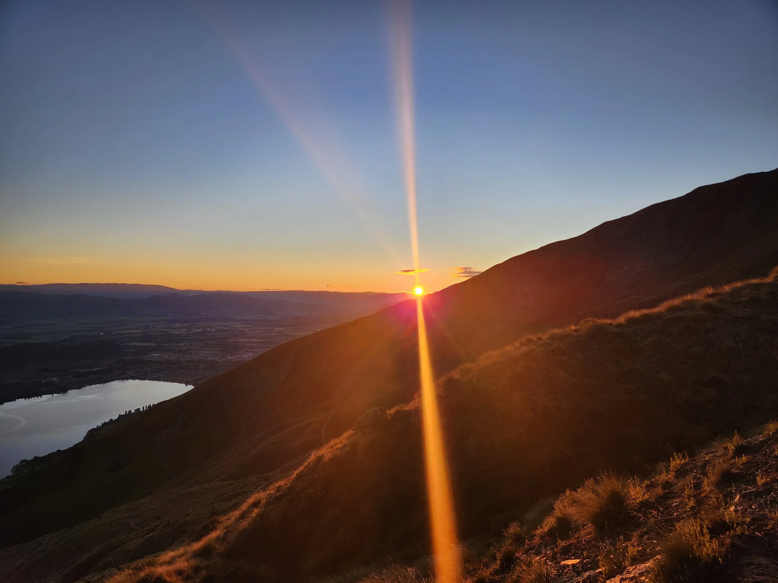 Sunset over a mountain with sunlight creating a vertical beam and a horizontal streak in the sky, overlooking a lake in a valley.