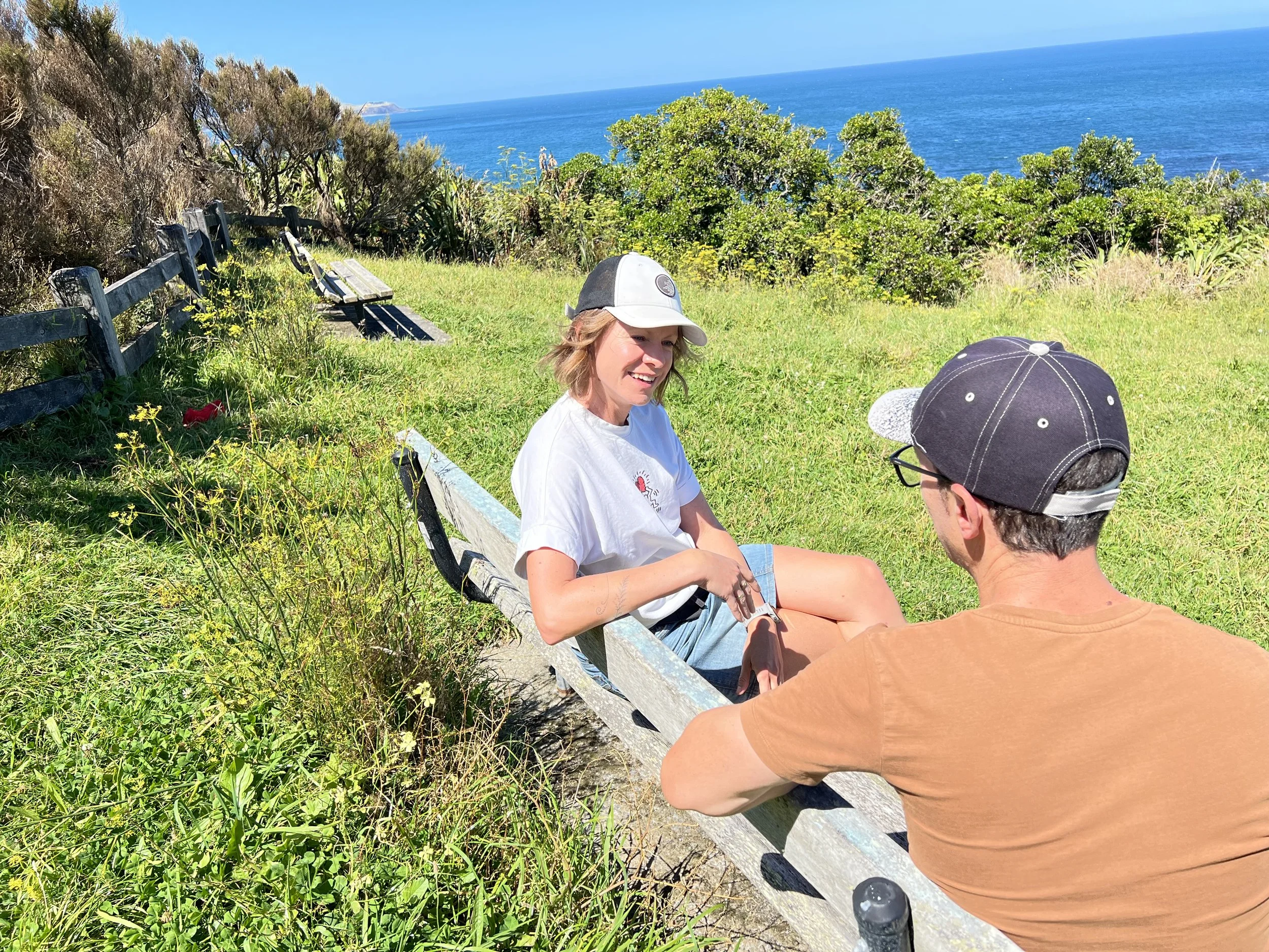 A young woman and a young man sitting on a park bench, smiling and talking, with the ocean and greenery in the background on a sunny day.