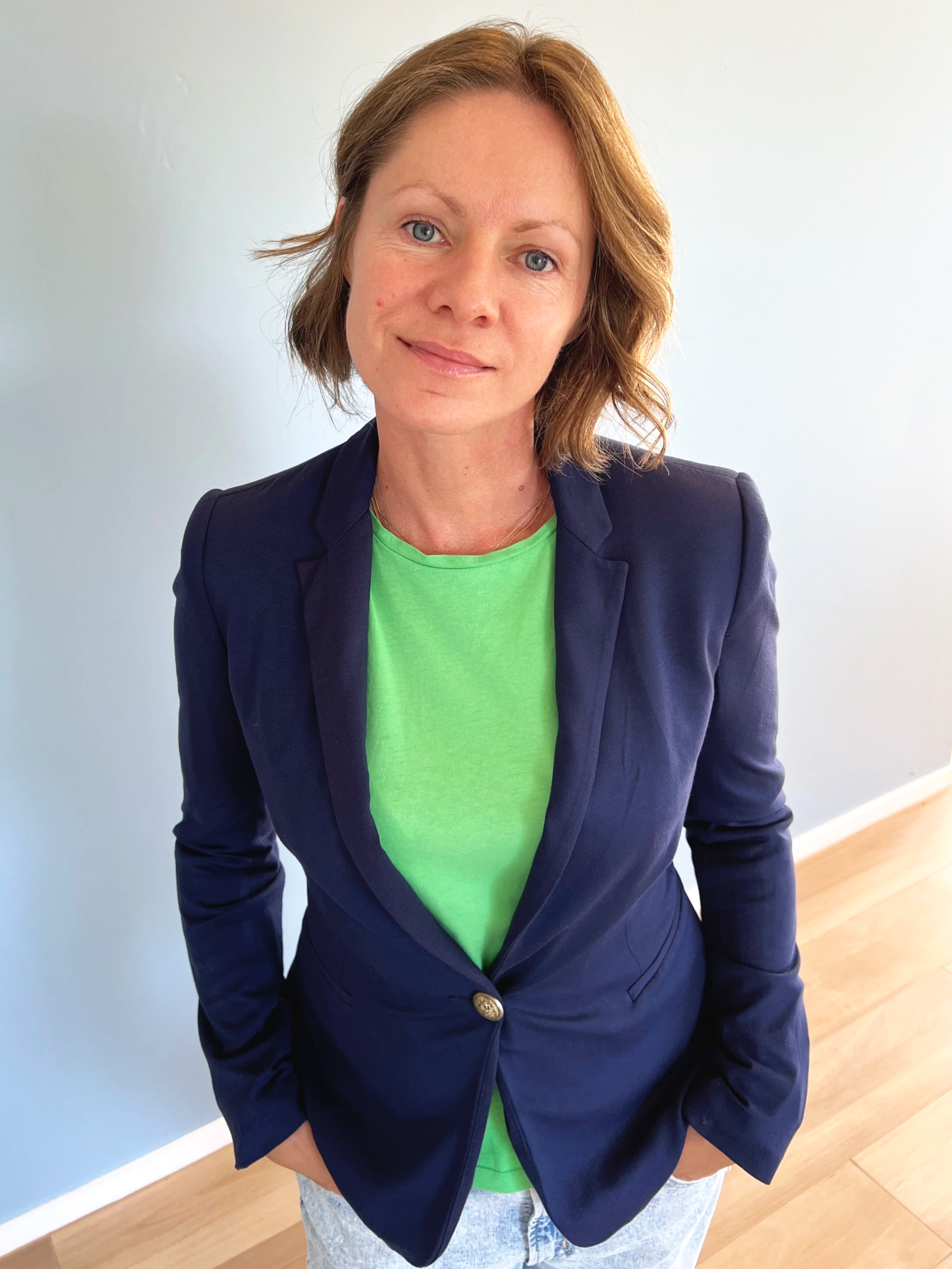 A woman with shoulder-length brown hair and blue eyes, wearing a navy blazer over a bright green t-shirt, standing indoors on a wooden floor against a plain light-colored wall.