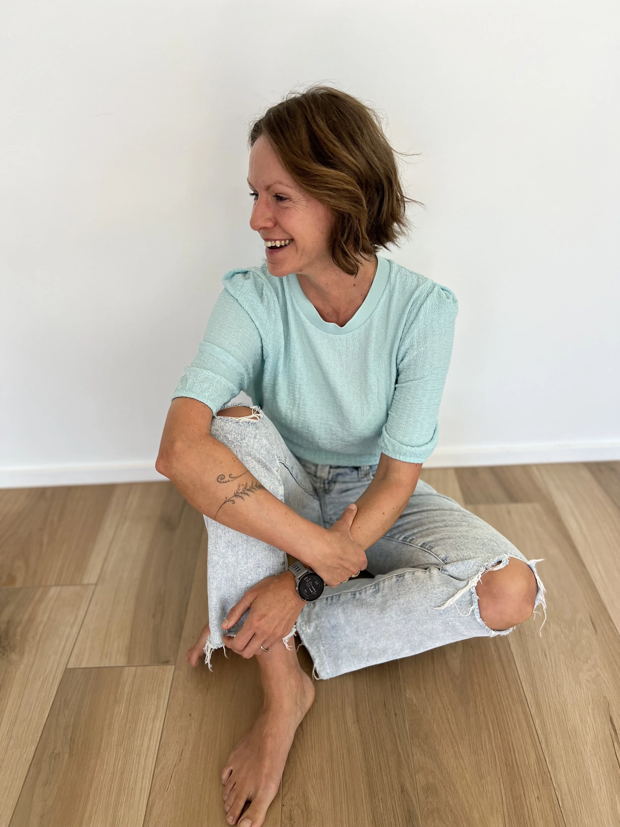 A woman with brown, shoulder-length hair, wearing a light blue top and ripped jeans, is sitting on a wooden floor, smiling and looking to her left.