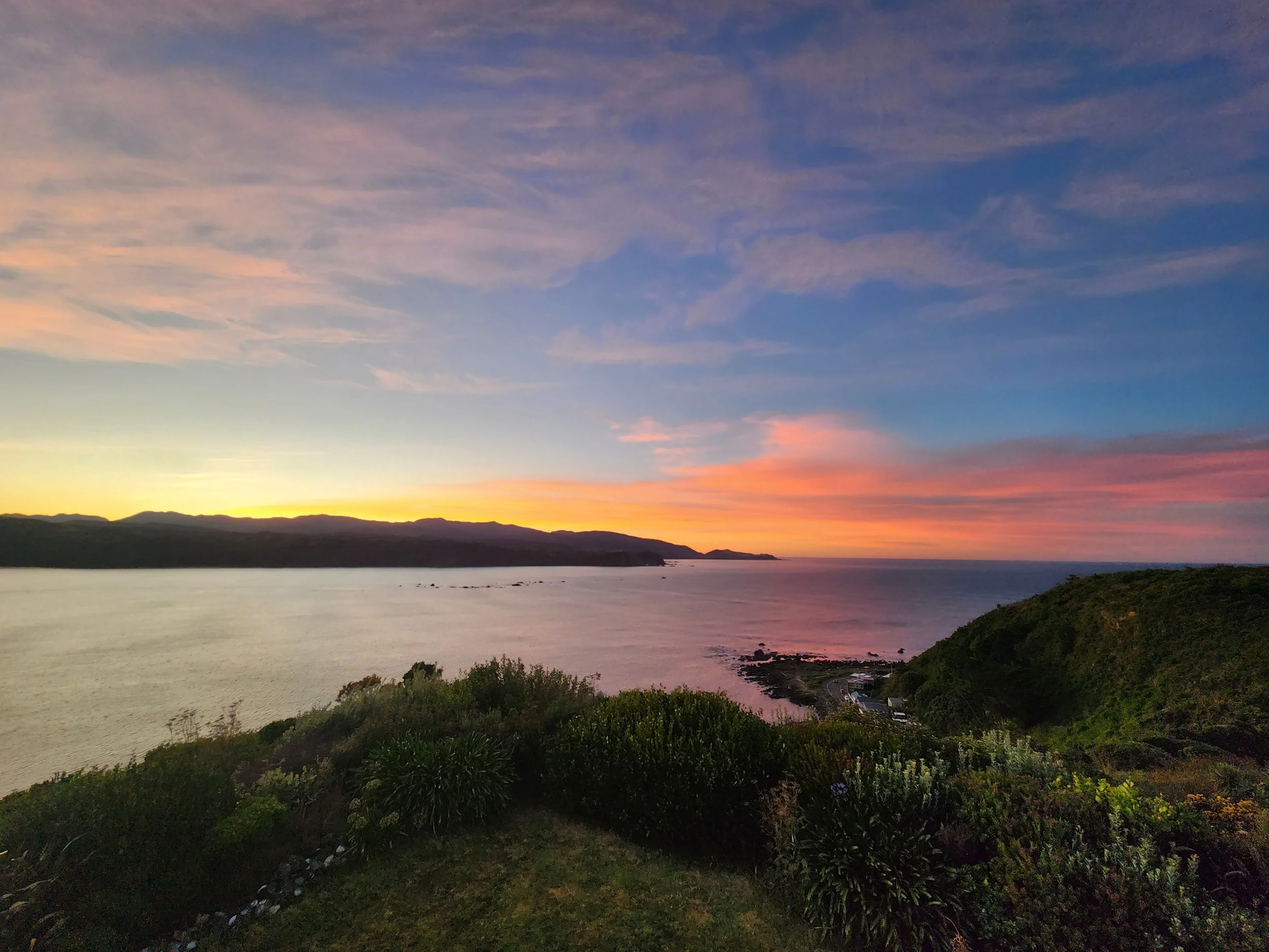 Sunset over the ocean with colorful sky, distant mountains, and green shrubbery in the foreground.