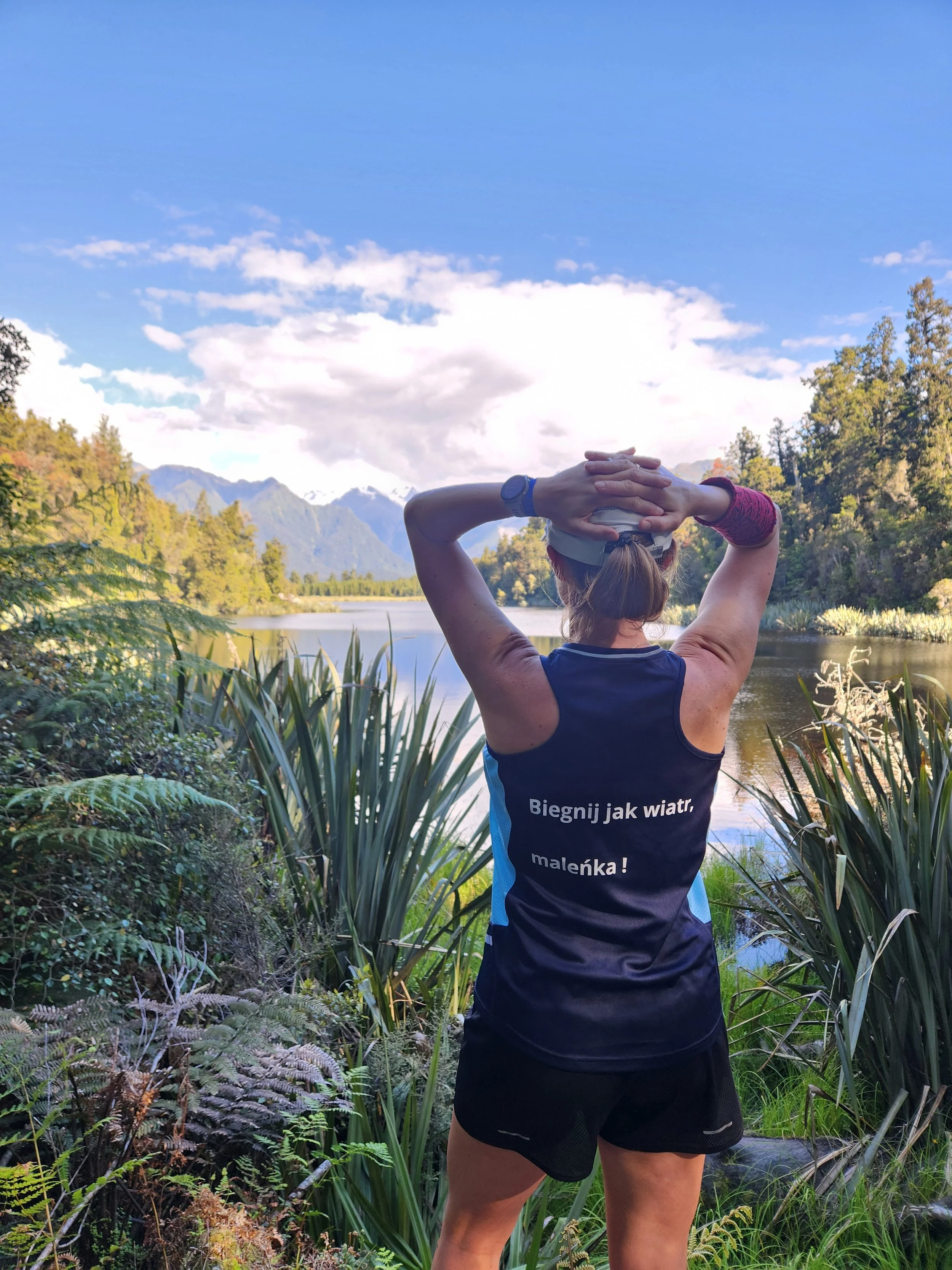 Athletic woman with a cap and wristwatch standing near a river, stretching her arms behind her head, with lush greenery and mountains in the background under a partly cloudy sky.