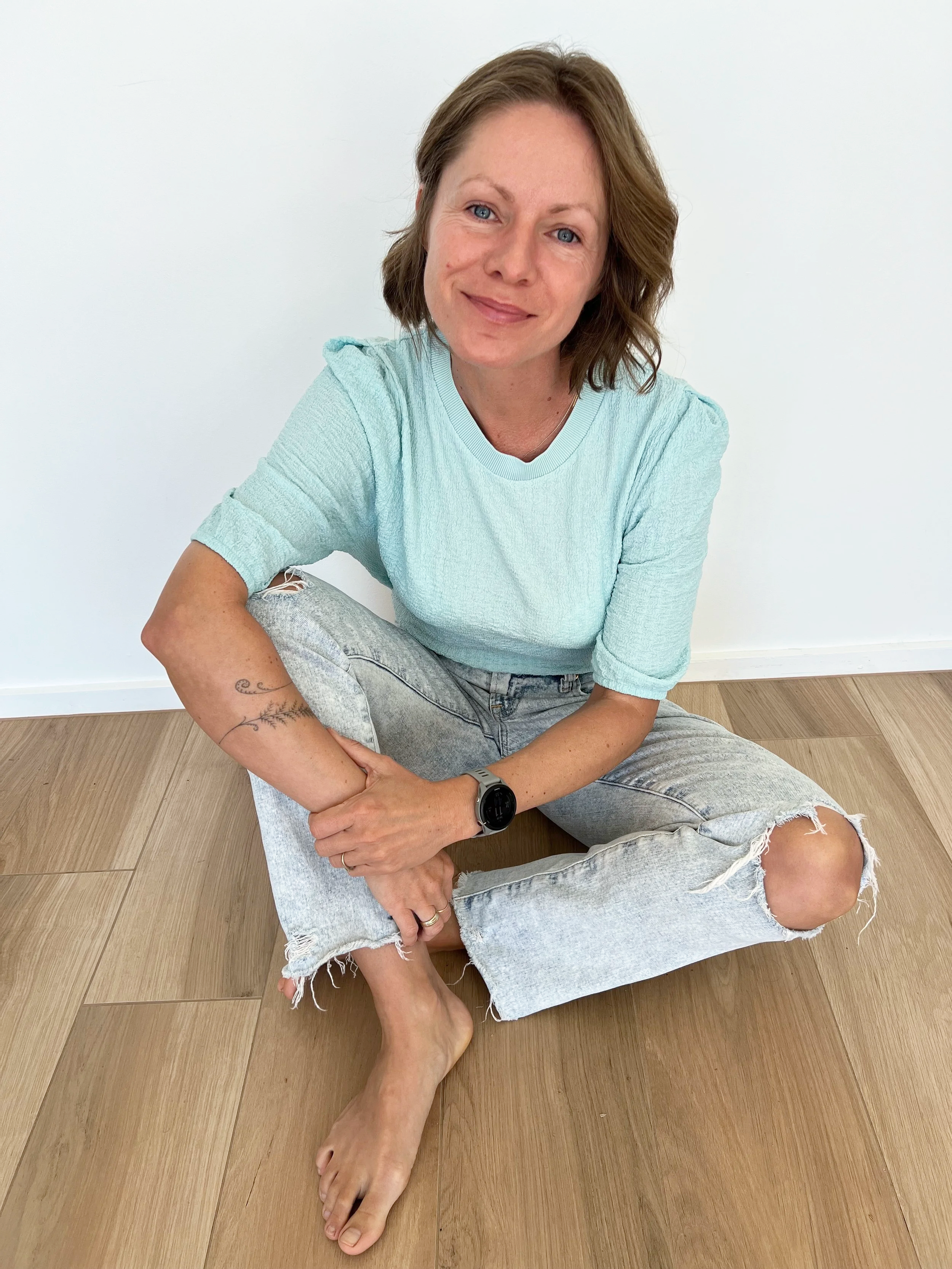 A woman with shoulder-length brown hair, blue eyes, and light skin, sitting on a wooden floor against a white wall. She is smiling, wearing a light blue t-shirt and ripped light-wash jeans, with a smartwatch on her left wrist. She has a tattoo on her right forearm and is barefoot.