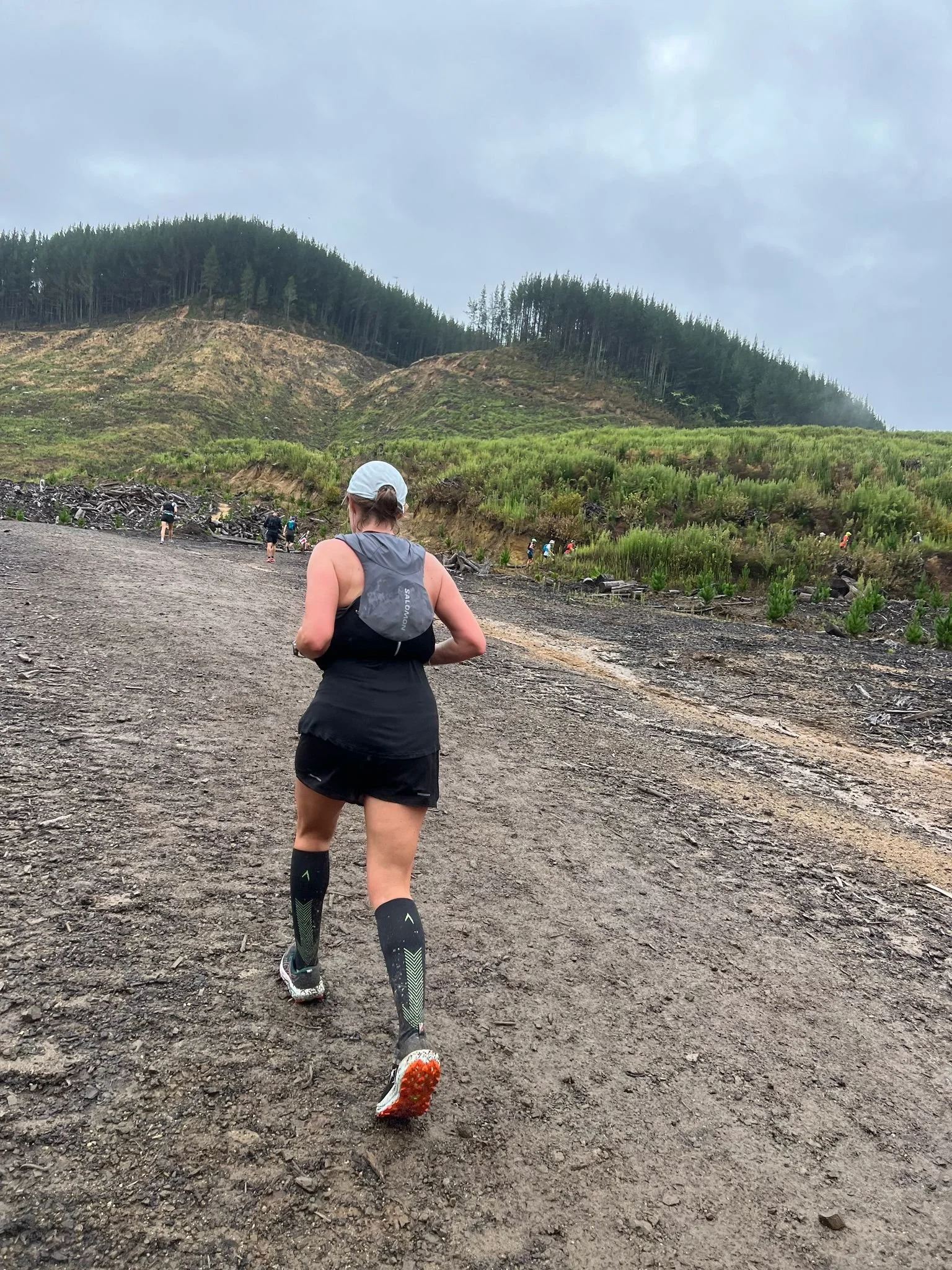 A woman in athletic gear running on a dirt trail in a mountainous outdoor setting with a cloudy sky.