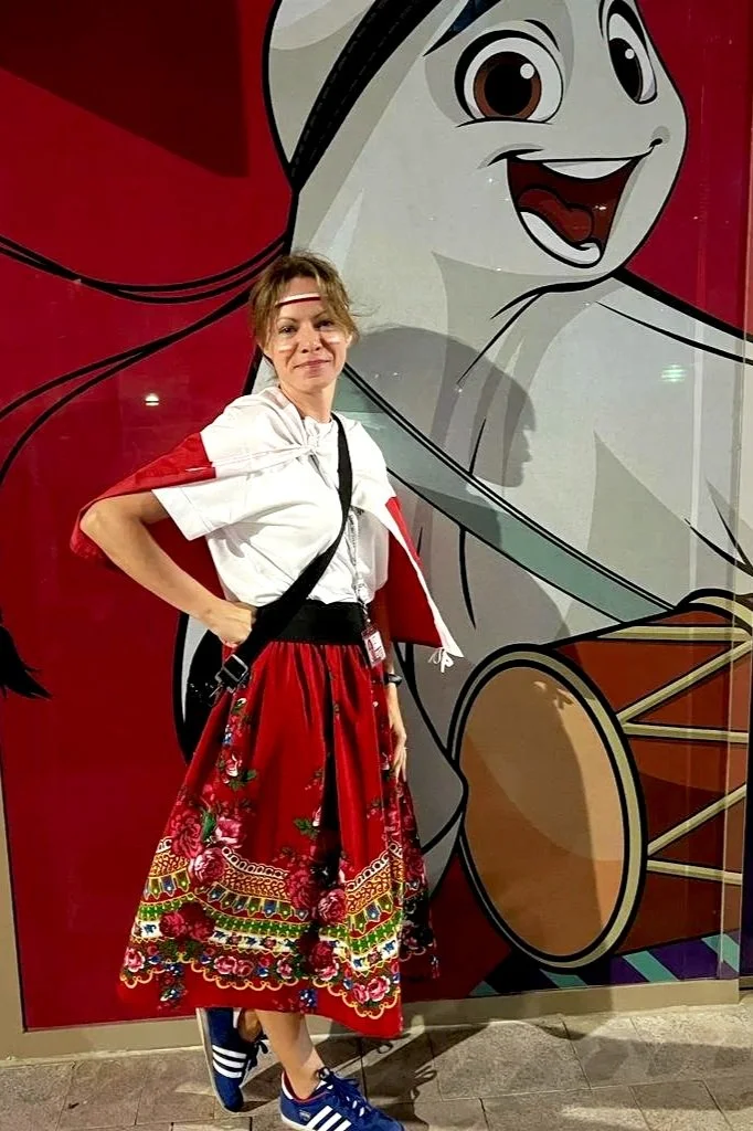 Woman standing in front of a large mural of a cartoon girl with white hair and brown eyes, wearing a red and black costume with embroidered floral and geometric patterns, blue Adidas sneakers with white stripes.