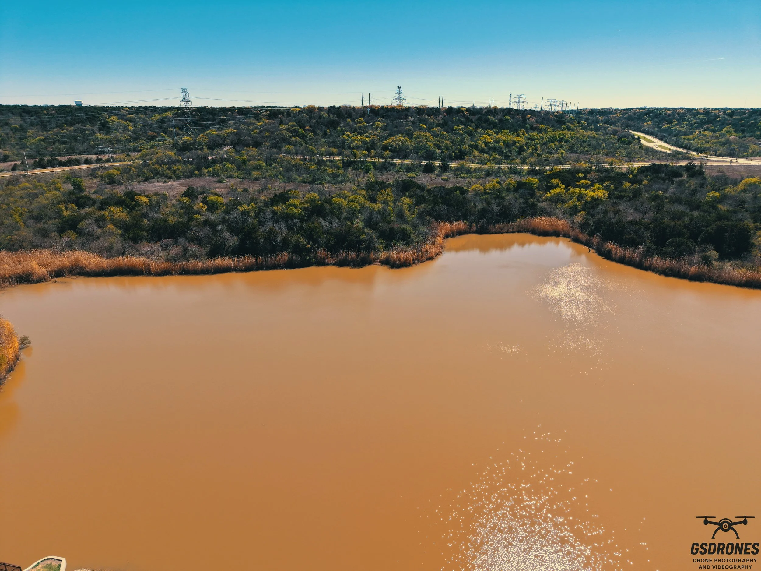 Aerial view of a muddy brown lake surrounded by trees and hills, with power lines and a highway in the background under a clear blue sky.
