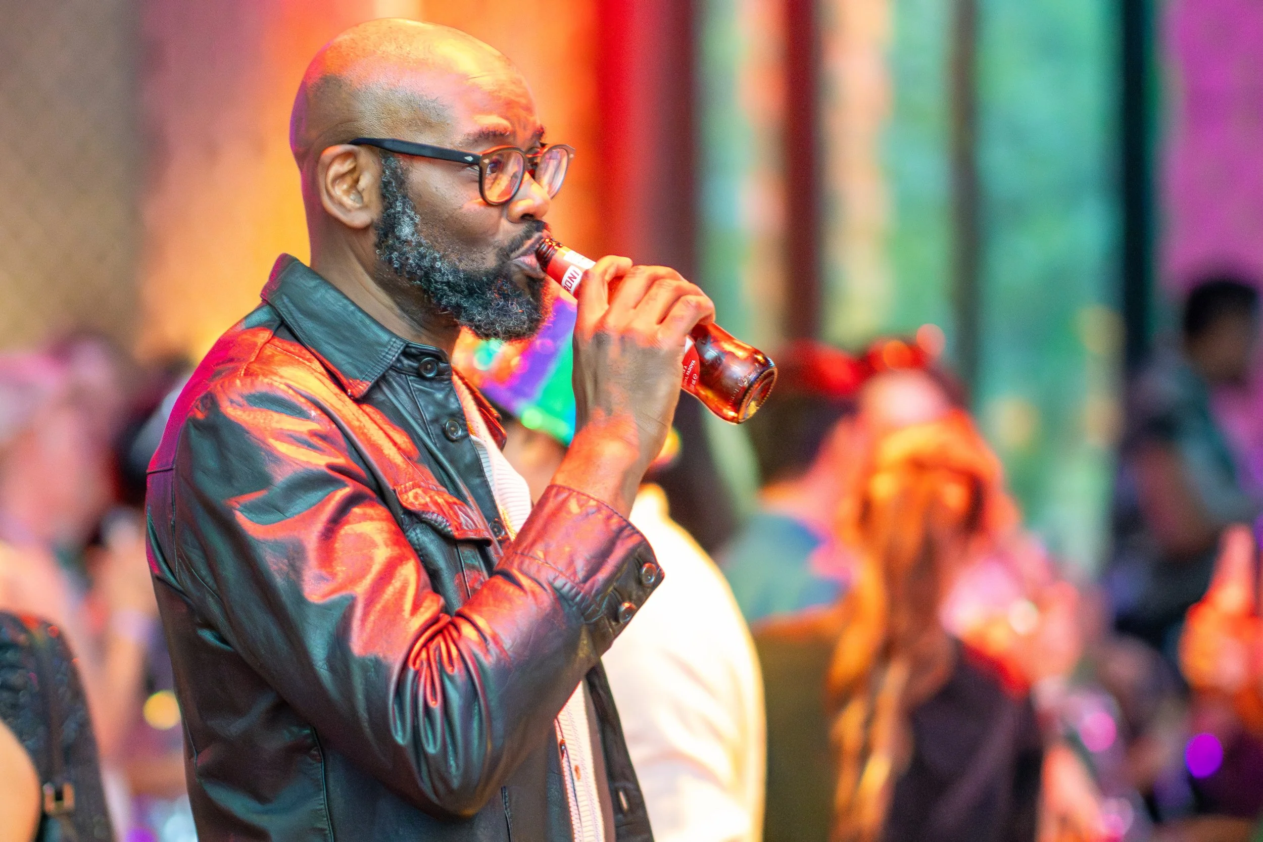 A man with glasses and a beard drinking from a bottle at an event with colorful background lighting.