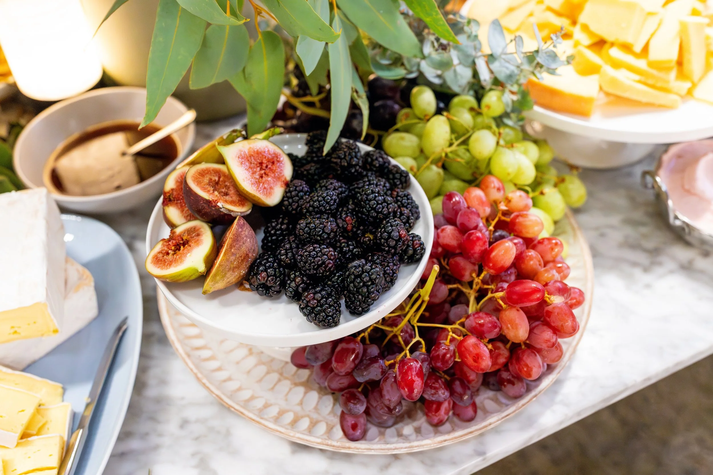 A platter with fresh figs cut open, blackberries, green grapes, and red grapes on a marble table, with cheese and a bowl of honey in the background.