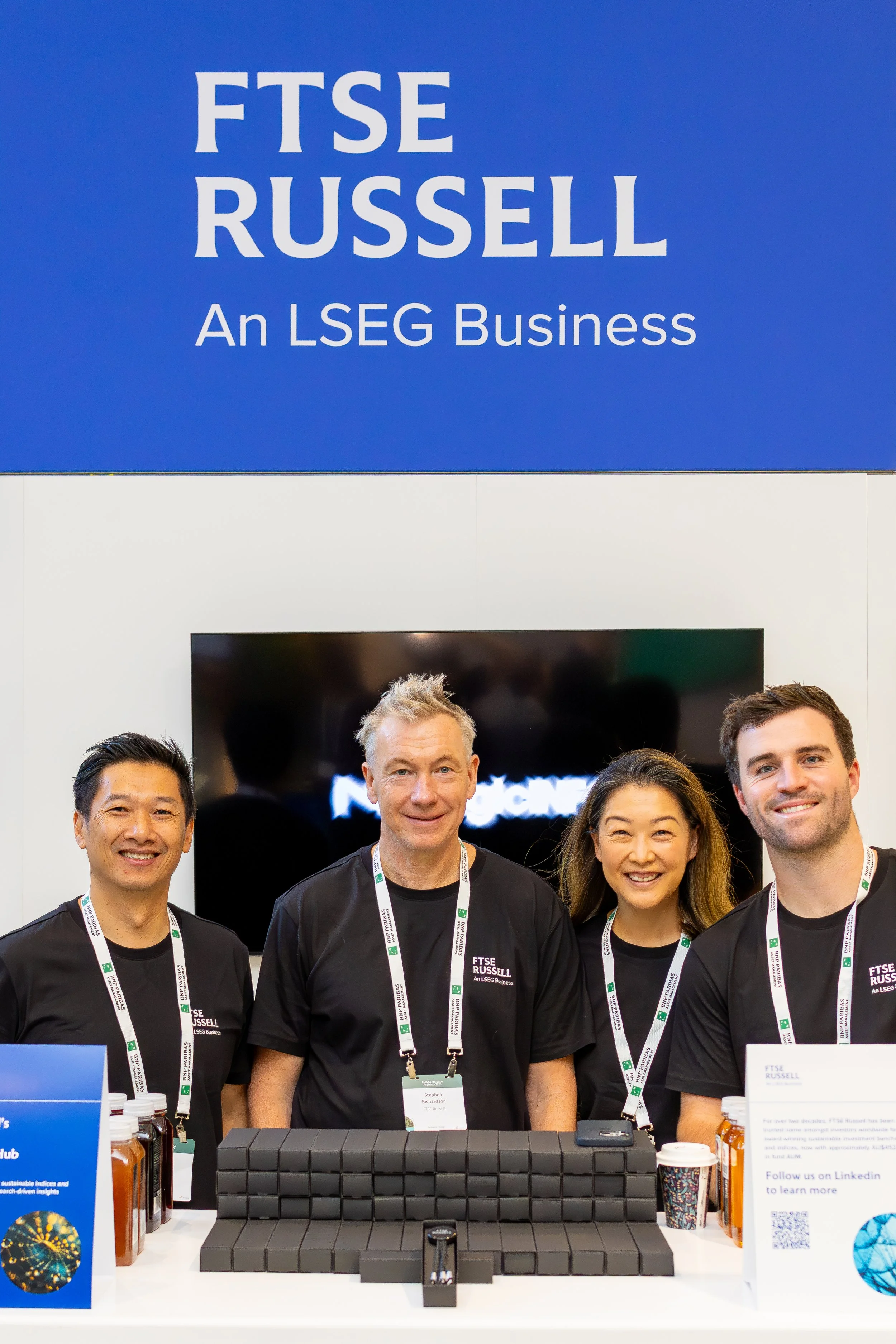 Four people smiling at a booth with a large sign that reads "FTSE RUSSELL An LSEG Business," with bottles and informational signs on the table.
