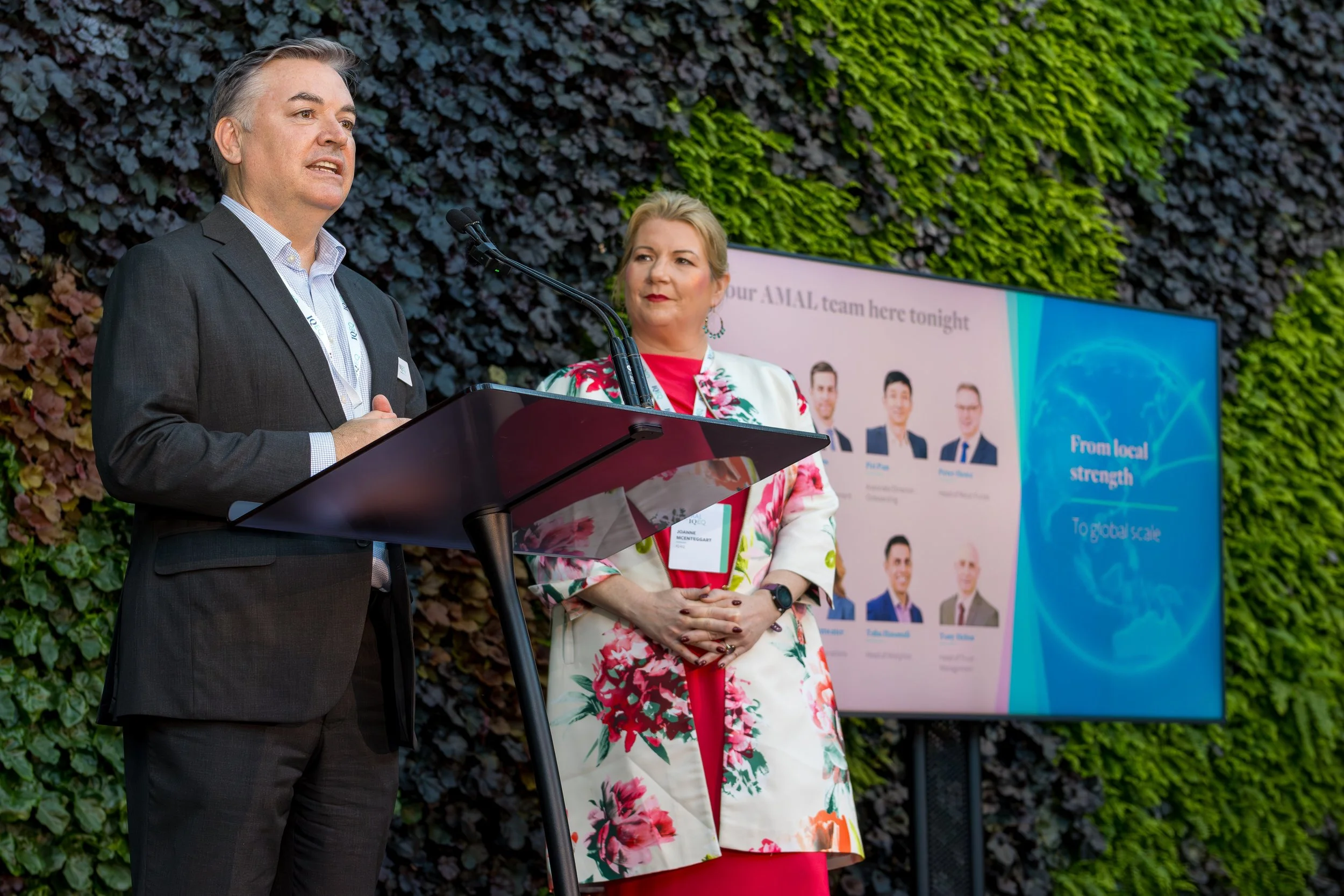 Two professionals speaking at a conference, one man in a gray suit and woman in a white floral blazer, with a large screen displaying a presentation behind them.