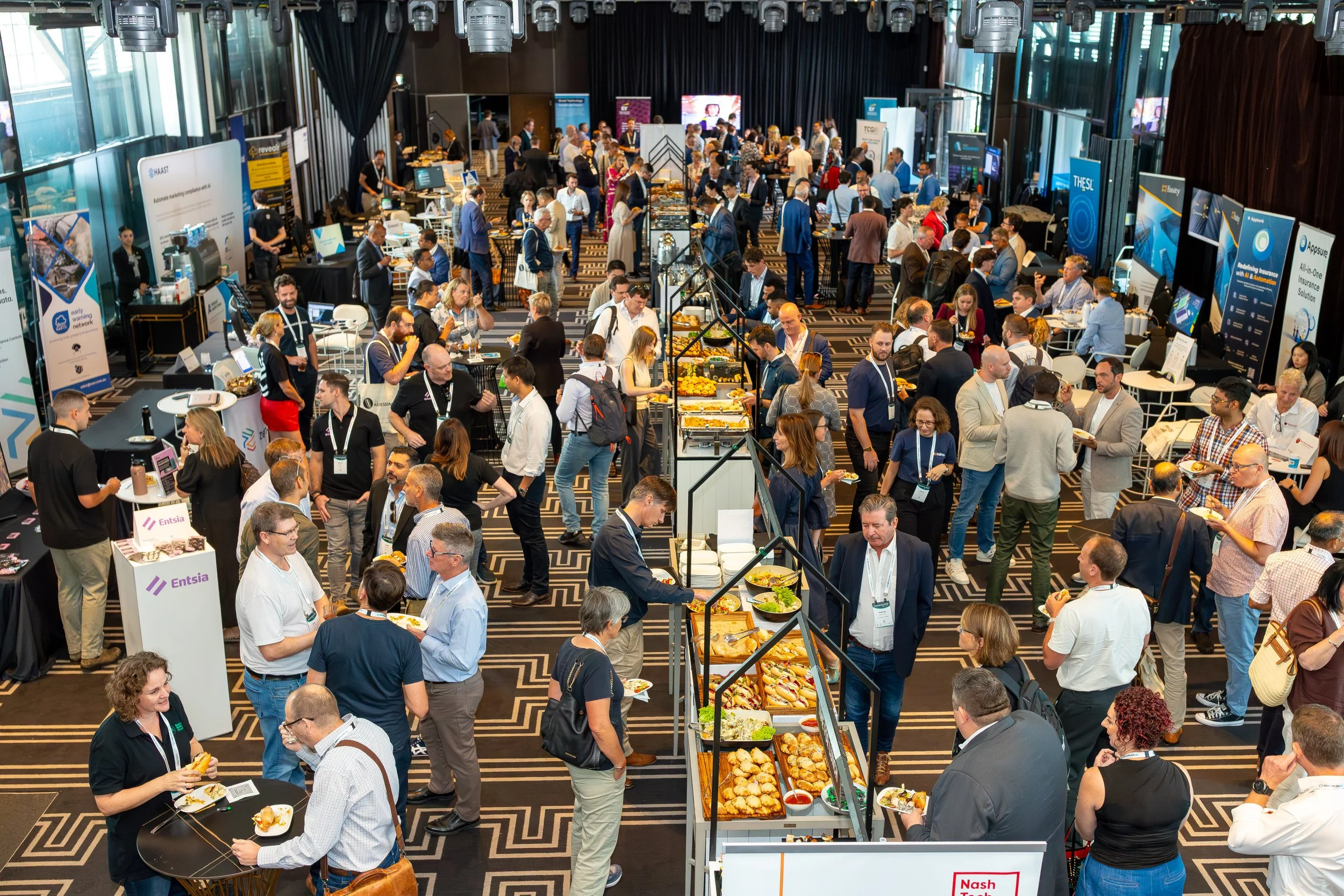 A busy conference or trade show with numerous booths, attendees, and buffet tables in a large, well-lit room with high ceilings and windows.