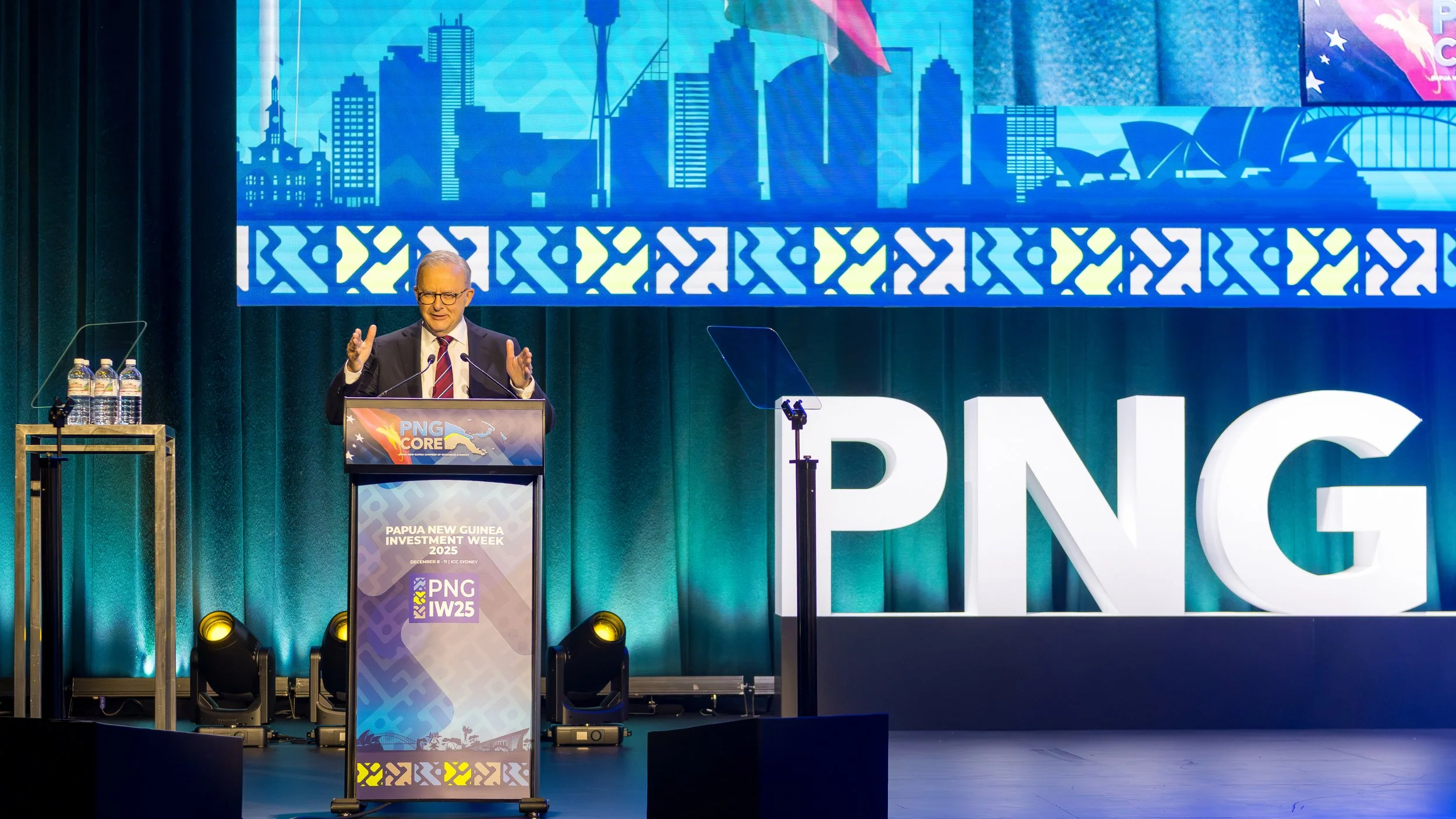 A man in a suit and glasses speaking at a podium during Papua New Guinea Investment Week 2025 event. The background displays the Sydney skyline and the Papua New Guinea flag, with large illuminated letters spelling PNG.