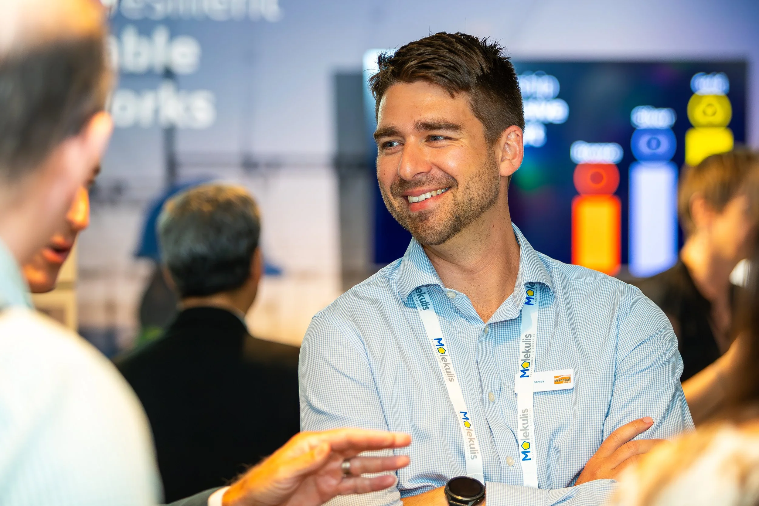 A man smiling and talking at a conference with a lanyard that says 'Molekuls' around his neck, others are in the background.
