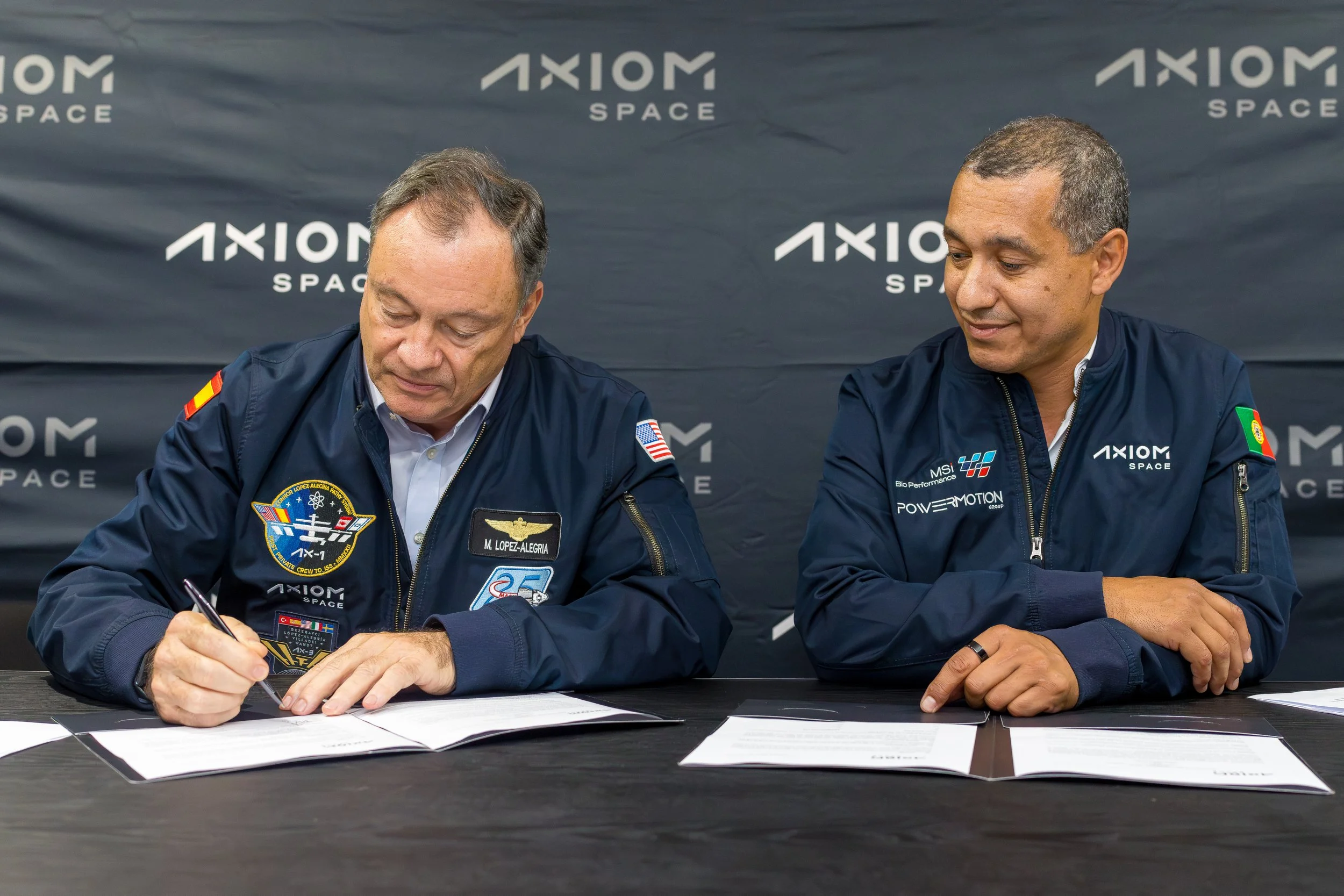 Two men in blue jackets sitting at a table, one signing a document while the other looks on, with a backdrop displaying the logo 'Axiom Space'.