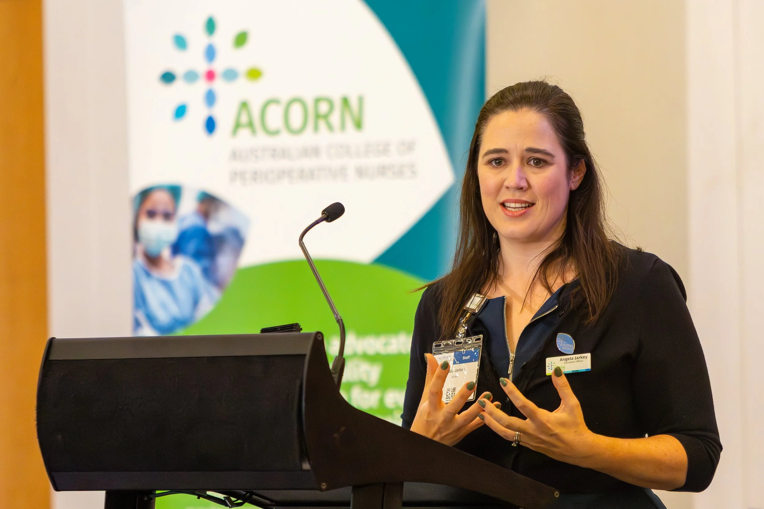 A woman with dark hair, wearing a black jacket and a name tag, is speaking at a podium with a microphone. Behind her is a banner for the Australian College of Perioperative Nurses (ACORN), showing images of healthcare professionals in masks.