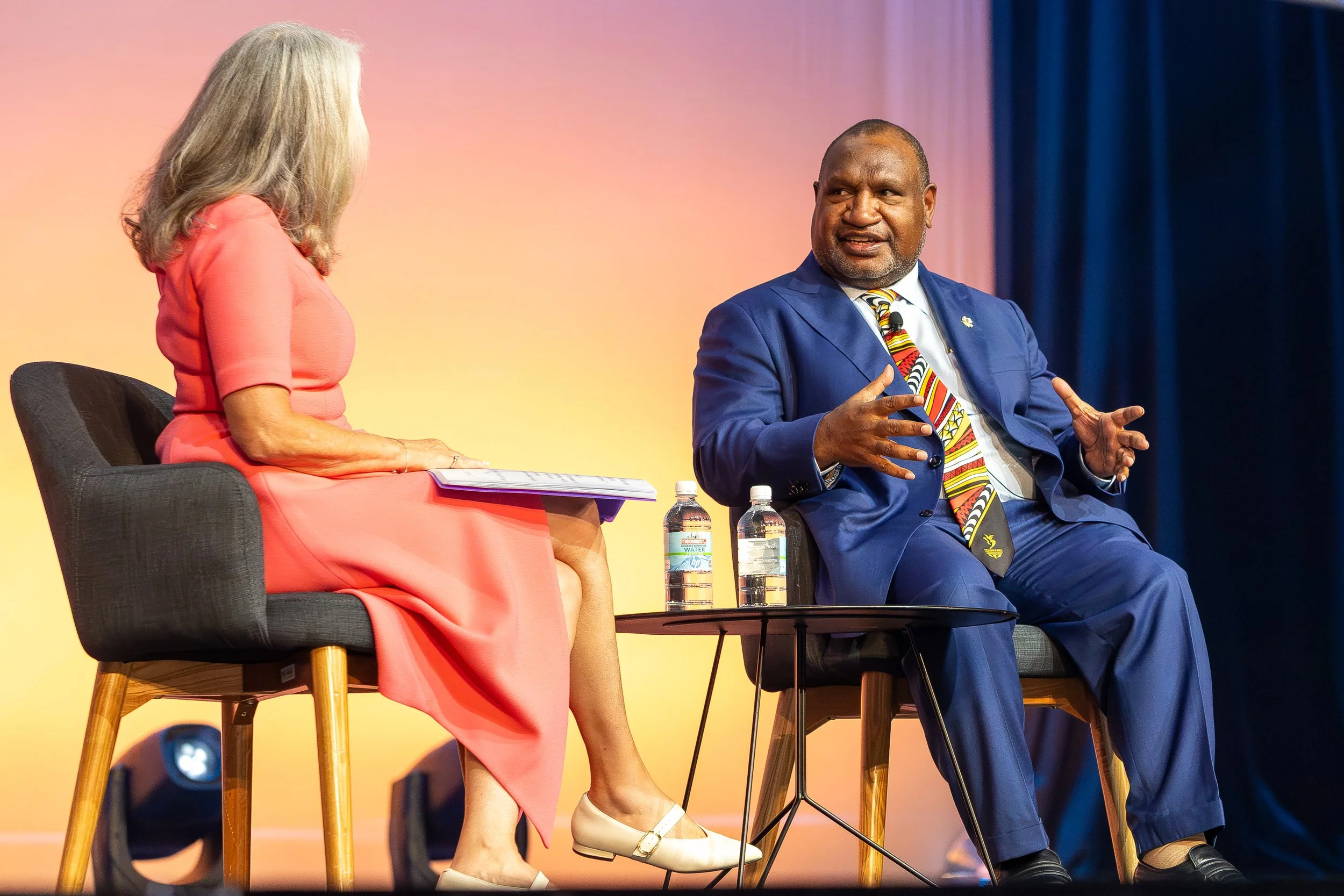 A woman in a coral dress and beige shoes sitting on a black chair, holding some papers and talking to a man in a blue suit with a colorful tie, sitting on a gray chair, with two water bottles on a small table between them, against a yellow-orange gra