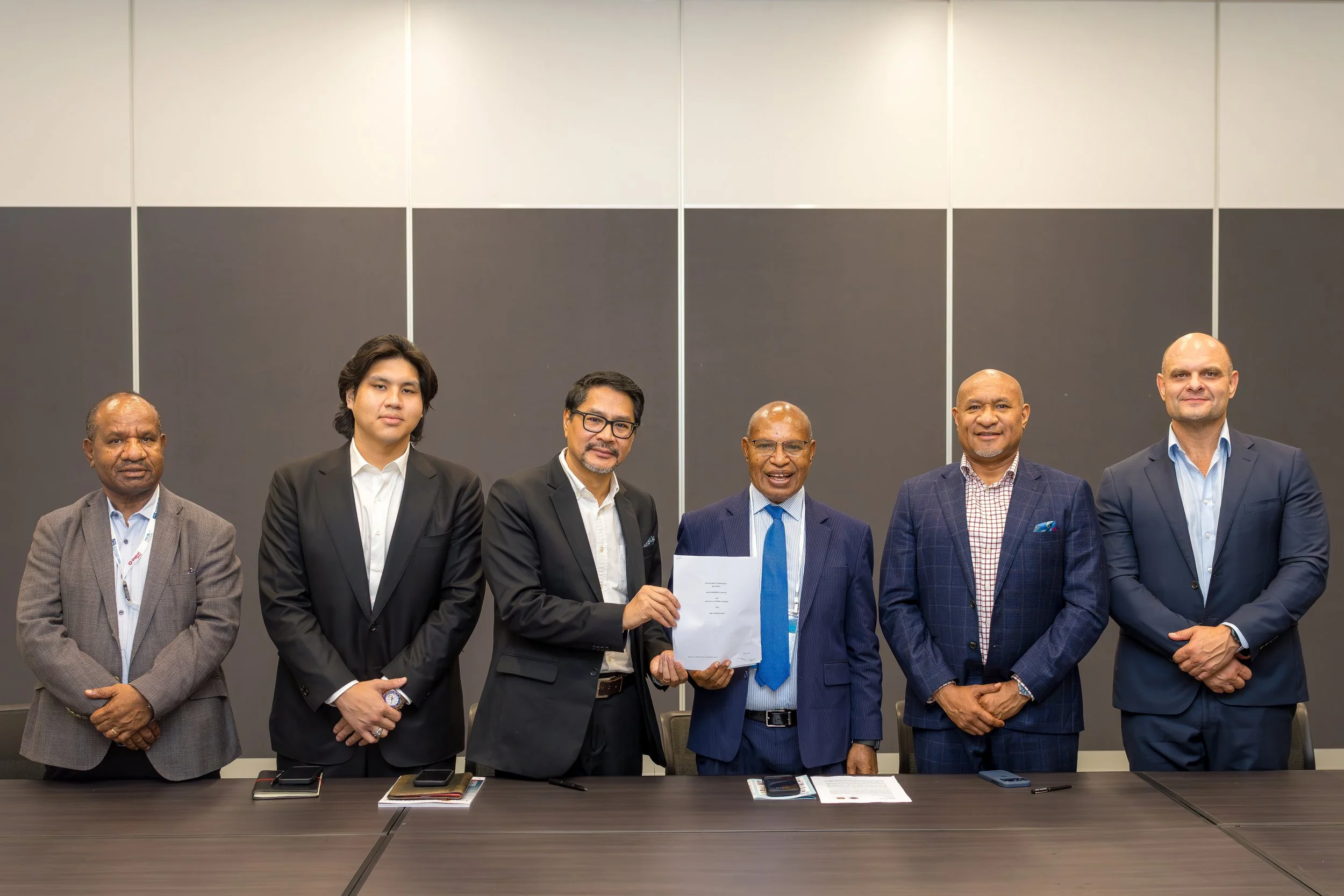 Seven men in formal business attire standing behind a conference table, one holding a document, in a modern conference room.