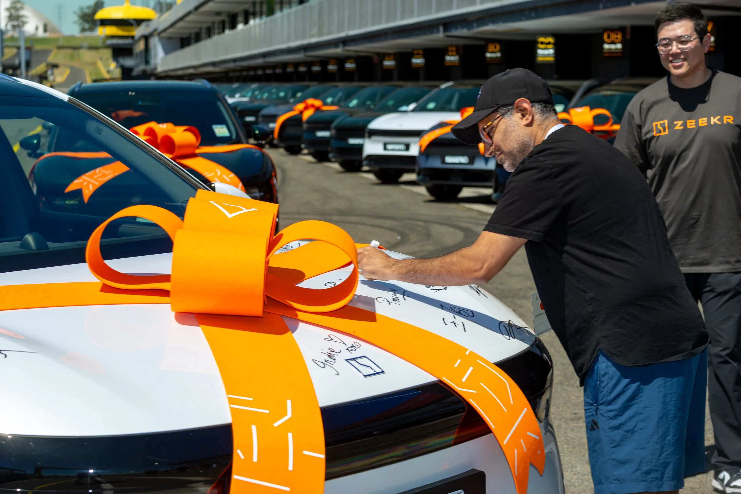 People at a car dealership or event with several new cars, some decorated with orange ribbons, and a man signing a document on the hood of one of the cars.