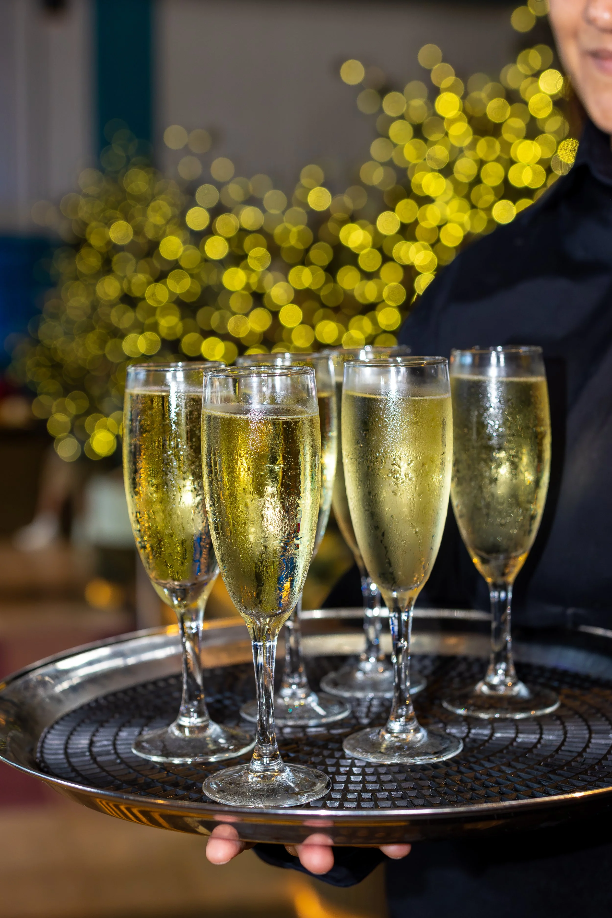 A person holding a tray with six glasses of champagne in front of a decorated Christmas tree with yellow lights.