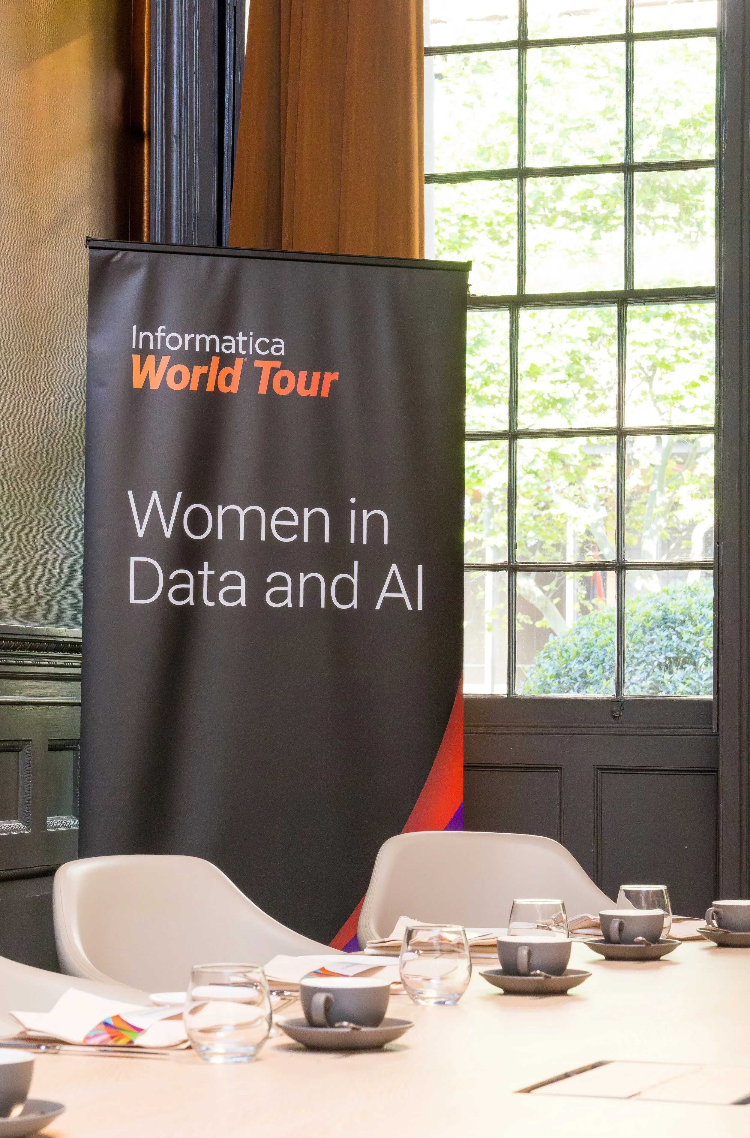 A banner with the title 'Women in Data and AI' at an event called Informatica World Tour, with a conference table set with cups, glasses, and napkins in a room with large windows and green foliage outside.