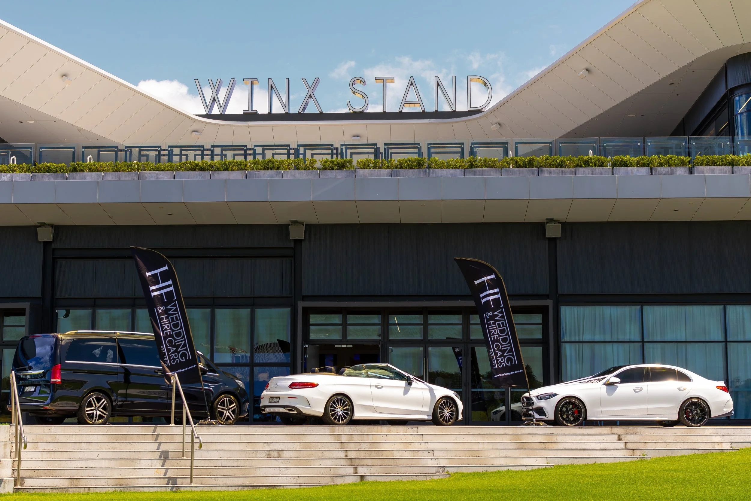 Multiple luxury cars displayed outside a modern building labeled 'WINX STAND' with flags promoting 'H Weddings & Hire Cars'.