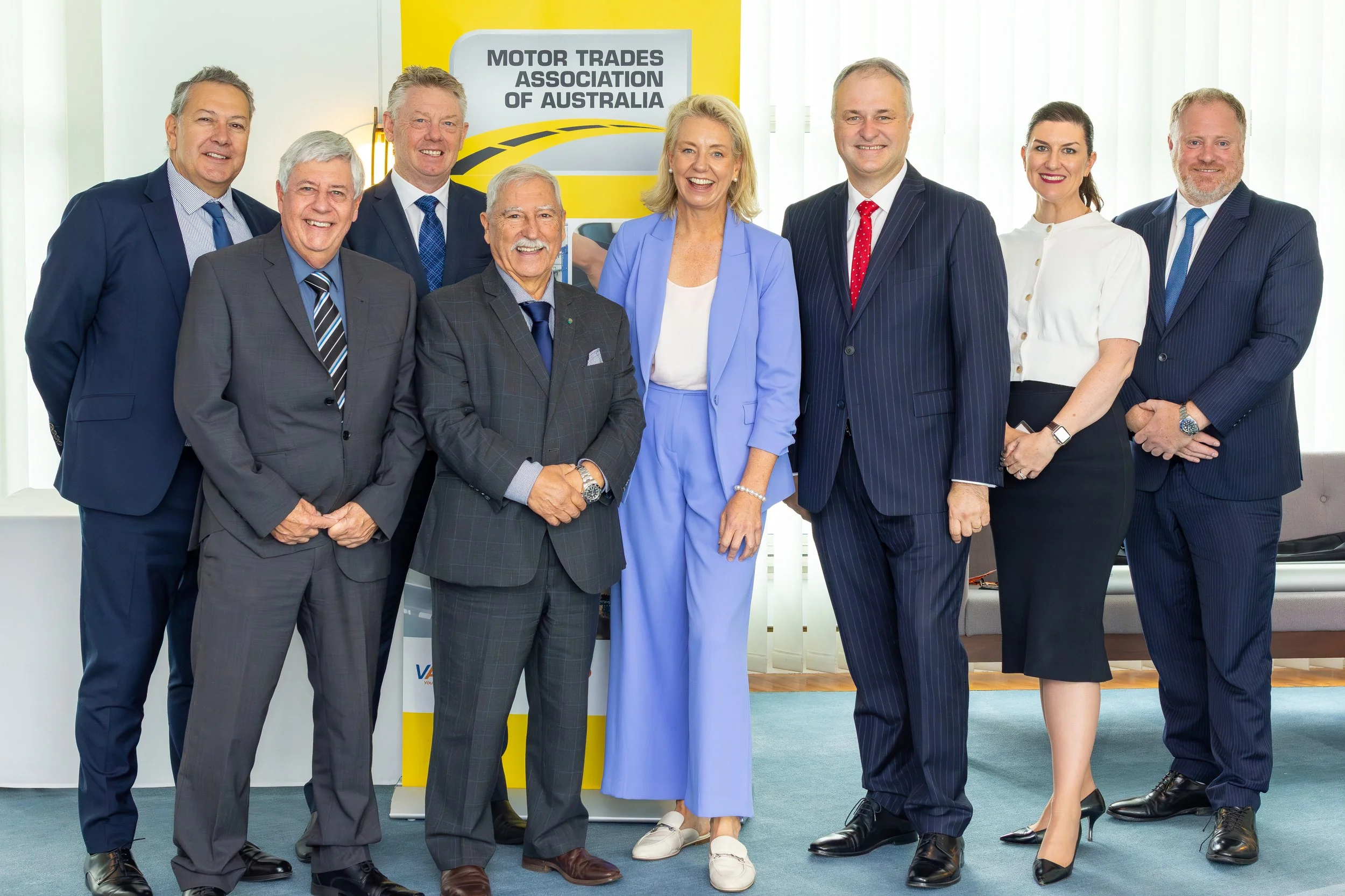 Group of nine business professionals standing in front of a banner for the Motor Trades Association of Australia, dressed in formal and business casual attire, smiling for the camera.