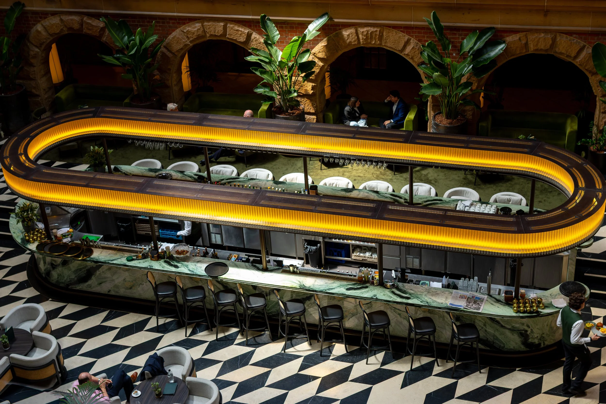 Interior view of a modern bar with a curved yellow-lit counter, high chairs, hanging glasses, and a marble countertop, with people seated in the background and a server walking by.