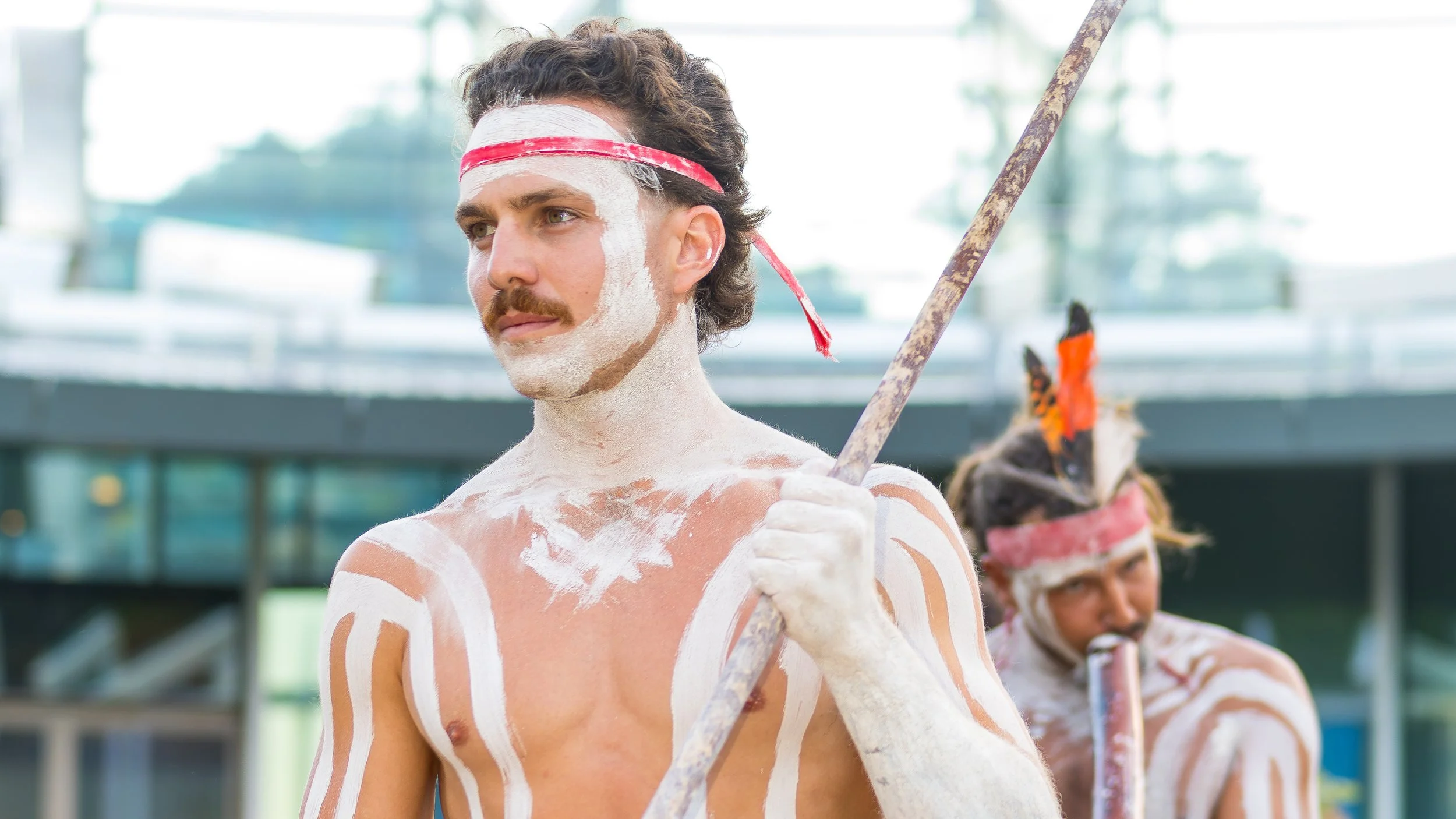 Two men with traditional body paint, face paint, and accessories, one holding a spear, in an outdoor setting with modern buildings in the background.