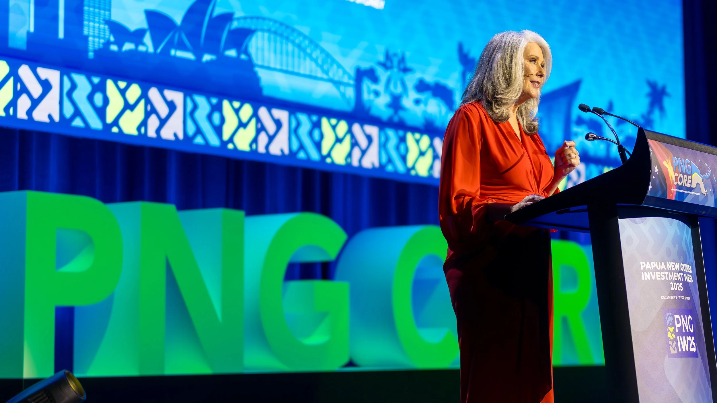 A woman with gray hair wearing a red dress giving a speech at a conference stage, with large green and blue letters spelling 'PNG' and a screen displaying a blue graphic background.