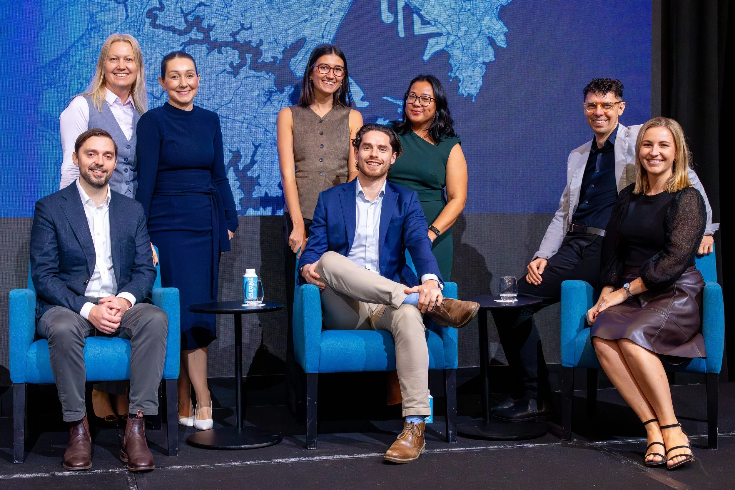 A group of nine diverse people, six women and three men, posing for a photo during a panel discussion or conference. They are standing and sitting in front of a large screen displaying a map of New York City.