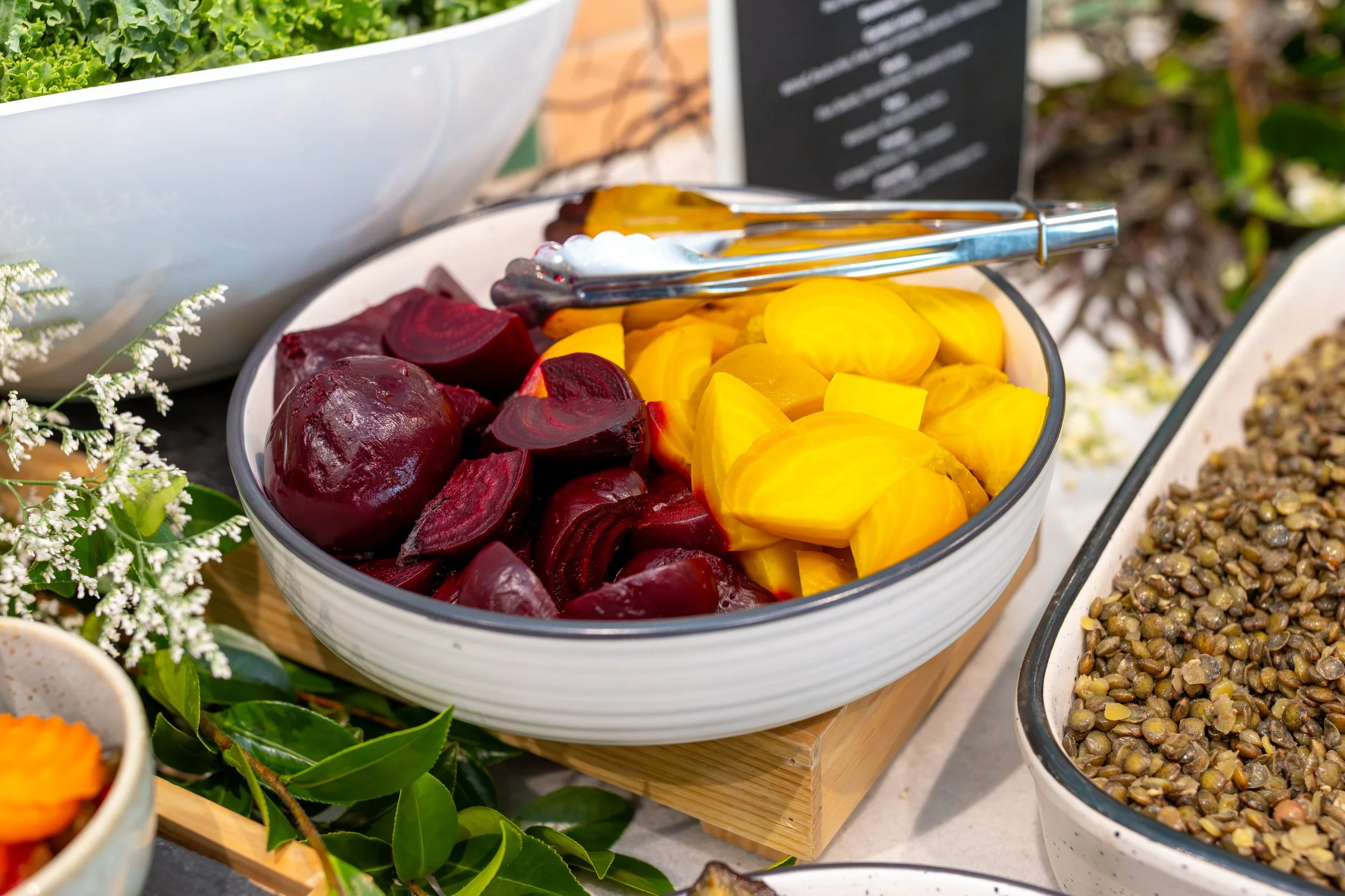 A bowl of sliced beets and yellow pickled vegetables at a buffet. Tongs rest on top of the bowl, and other dishes are partially visible nearby.