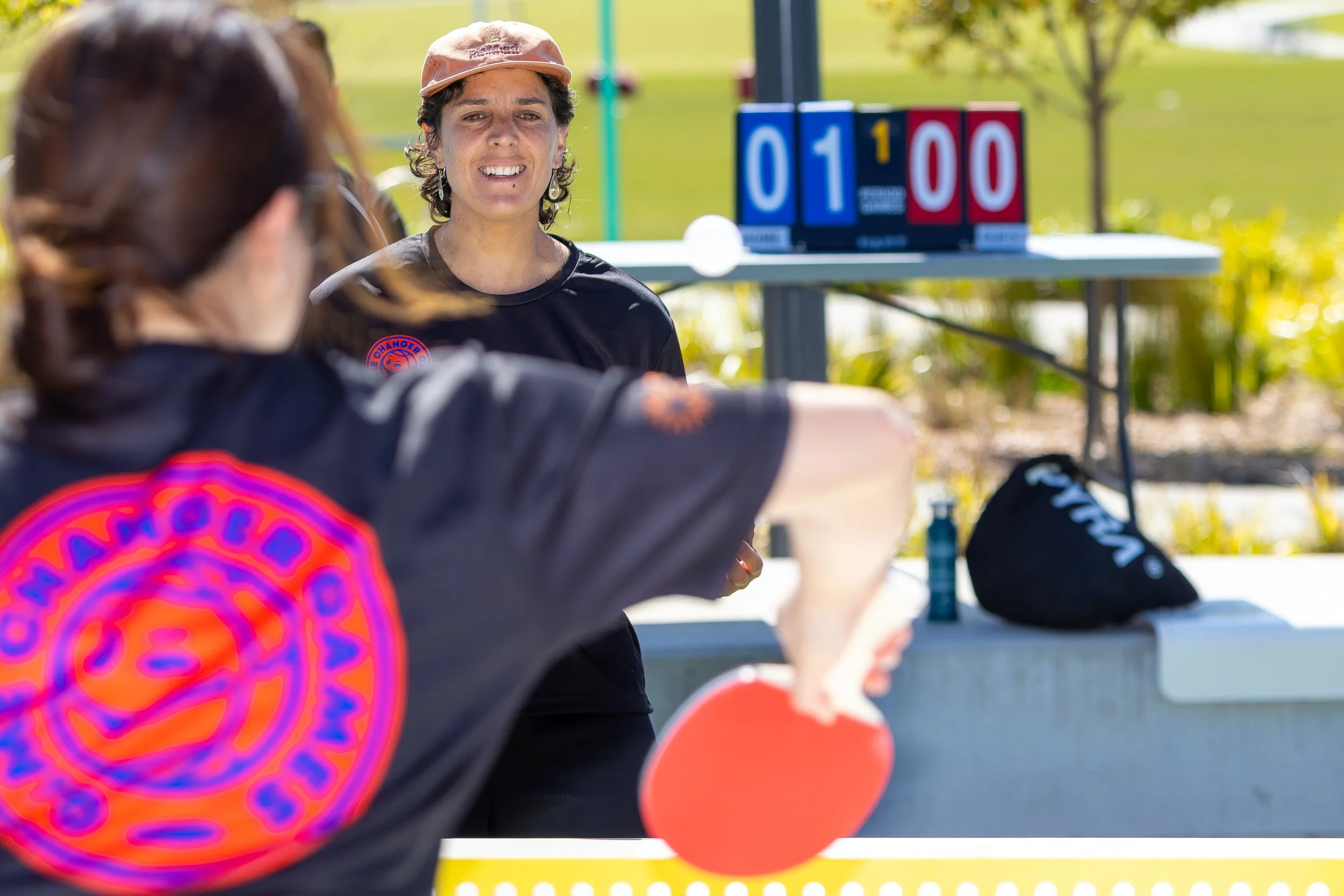 Two women playing pickleball outdoors, with a scoreboard showing 1 to 0 in the background, on a sunny day.