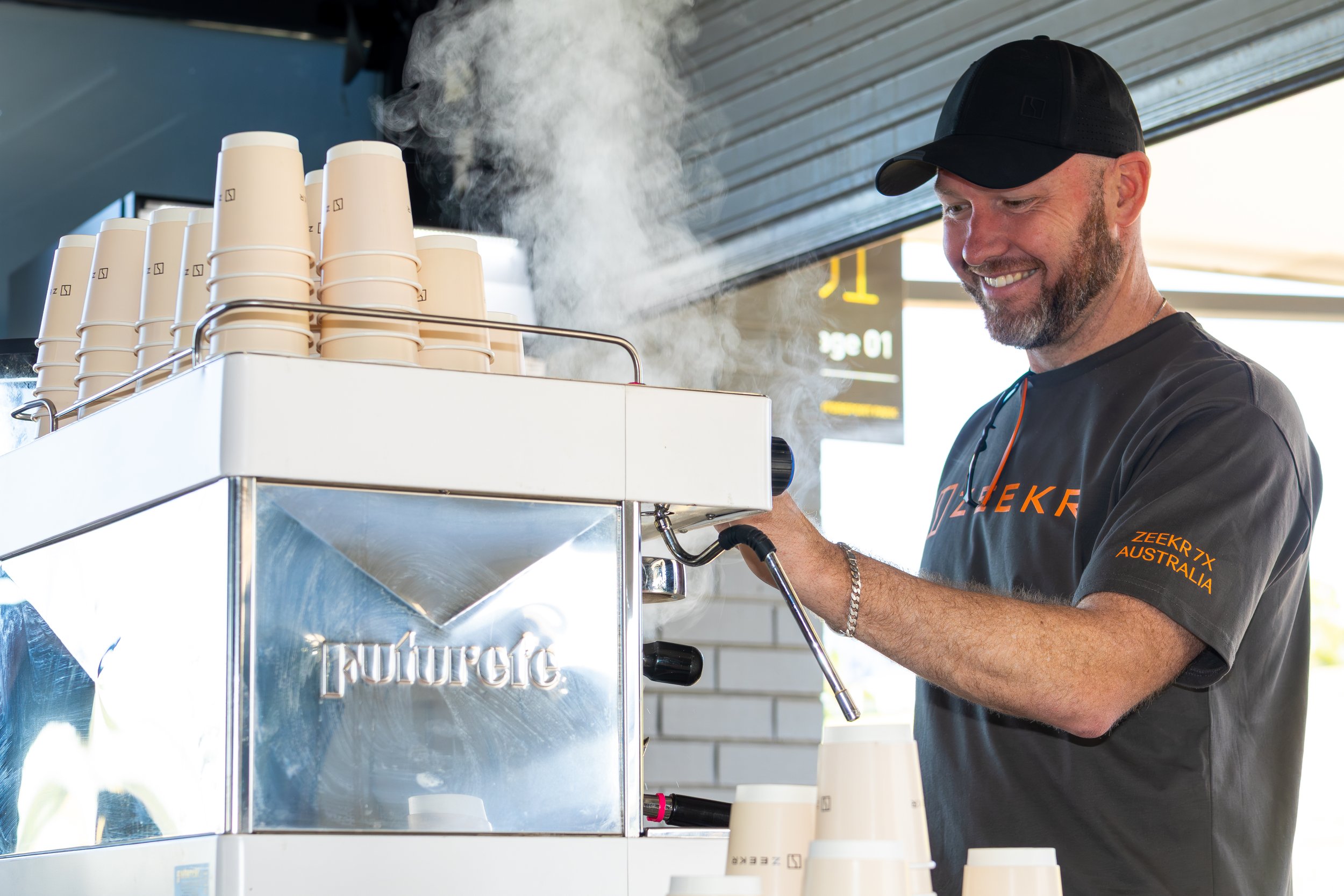 A man smiling and operating a coffee machine at a coffee shop, with the machine producing steam and disposable cups stacked on top.