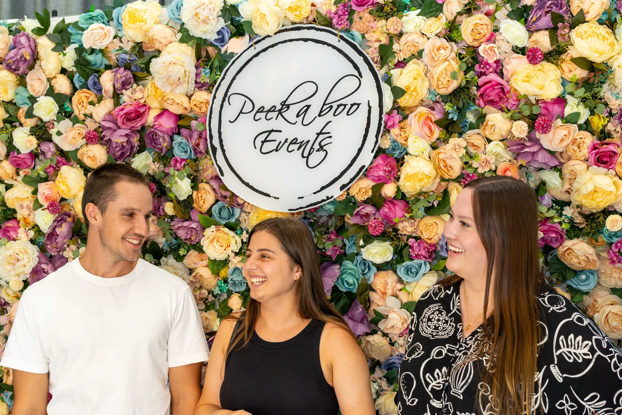 Three people smiling and talking in front of a colorful floral backdrop with a circular sign that reads 'Peeka boo Events'
