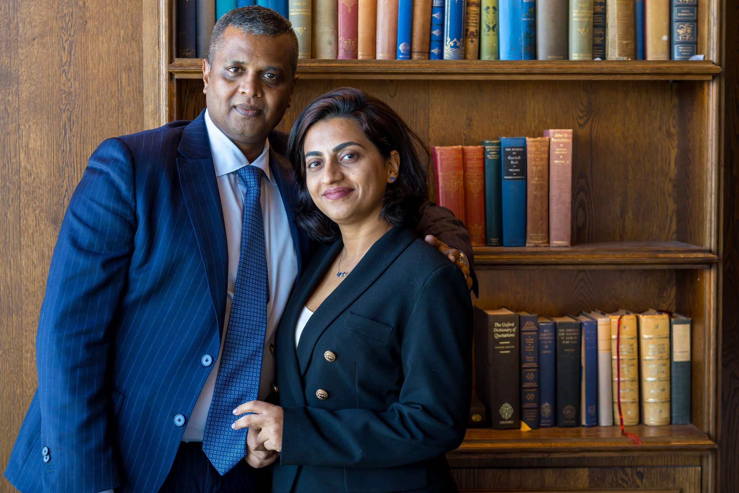 A professional man and woman standing together in front of a bookshelf, looking at the camera. The man is wearing a blue pinstripe suit and the woman is dressed in a black blazer.