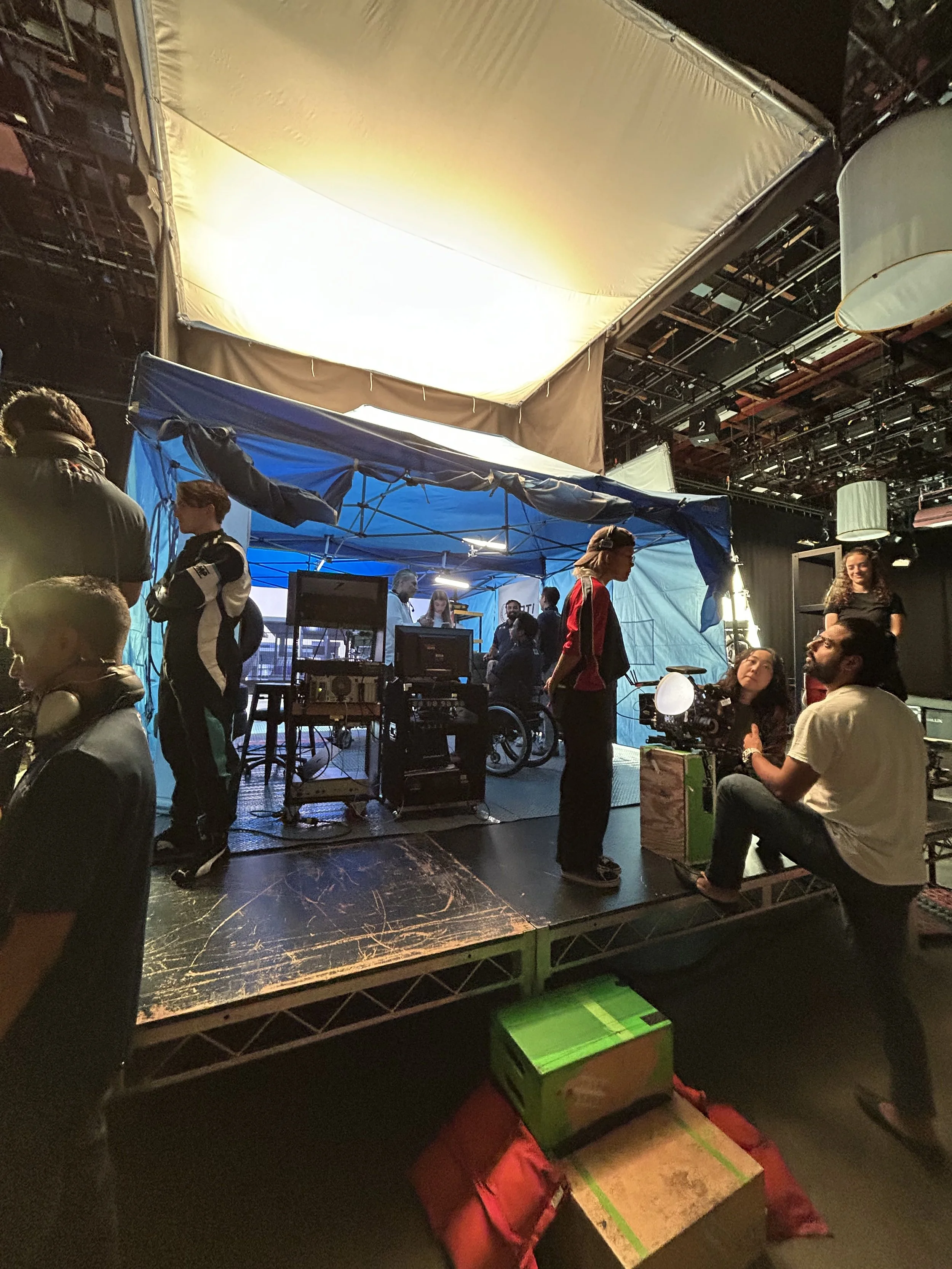 People gathered around a blue tent with electronic equipment, some seated and some standing, in a dimly lit indoor setting with stage lighting and hanging lamps.