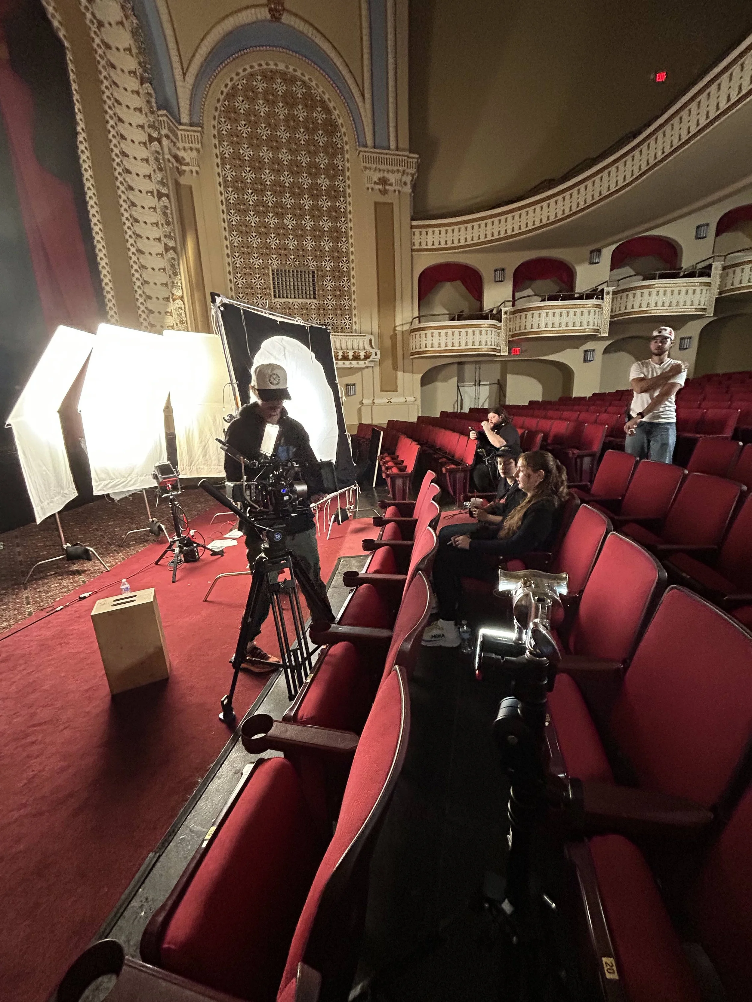 Interior of a theater with red seats, a camera crew setting up equipment for filming, and a few people sitting and standing, with ornate architectural details and balconies.