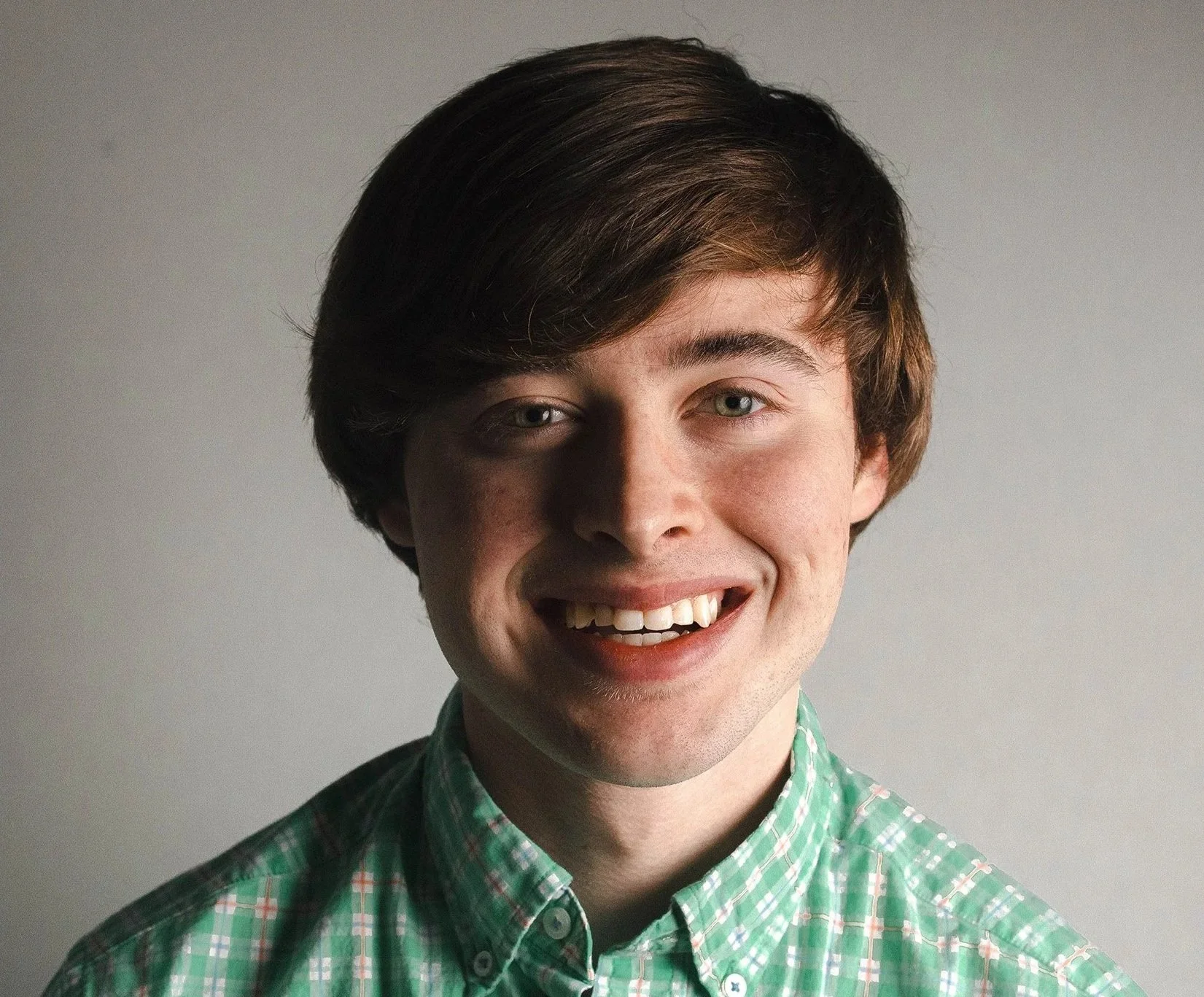 Portrait of a young man with dark brown hair, light eyes, smiling, wearing a green and white checkered shirt, against a plain background.