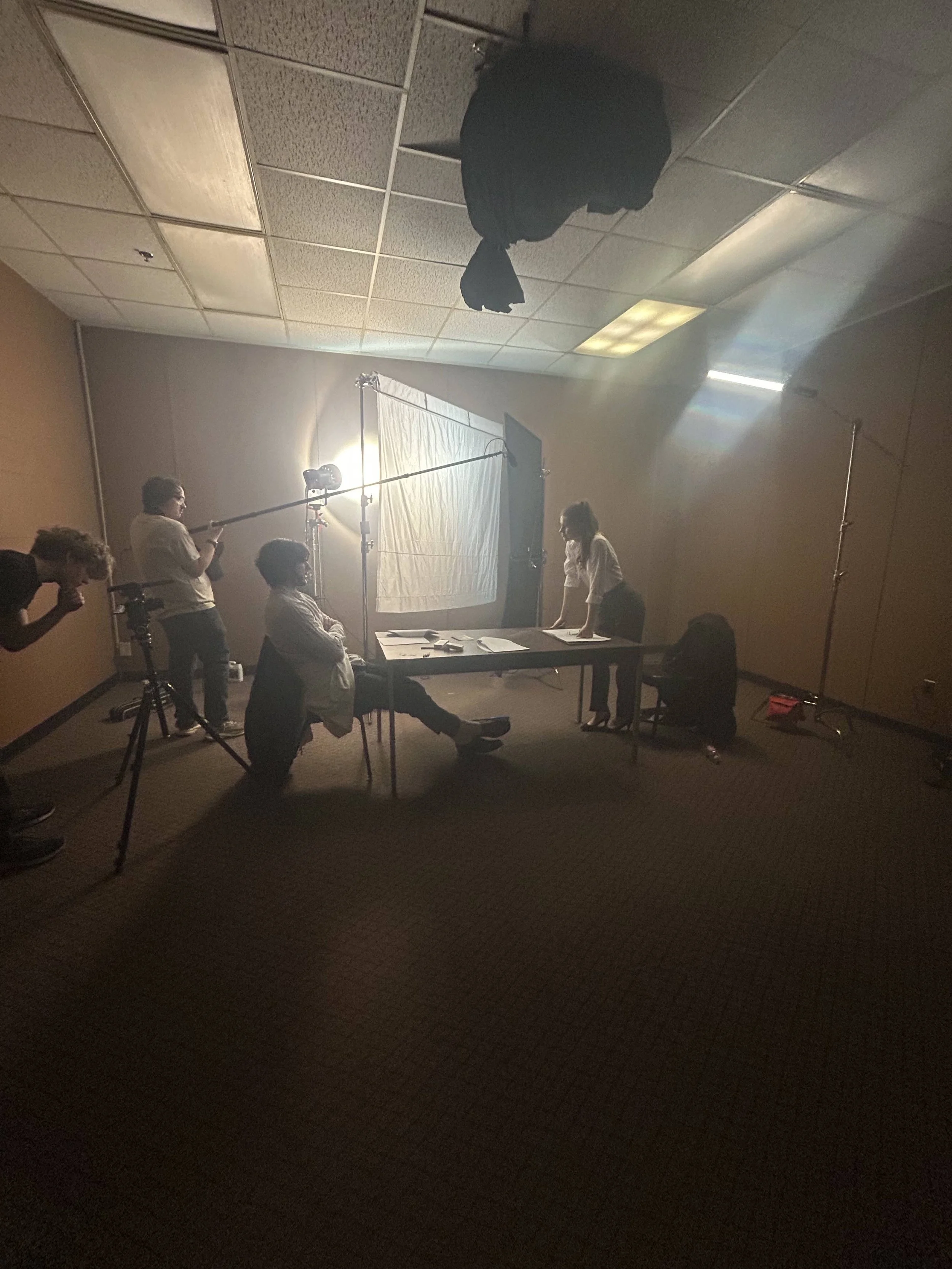 Video production set with lighting, a white backdrop, and people working with camera and equipment in a dimly lit room.