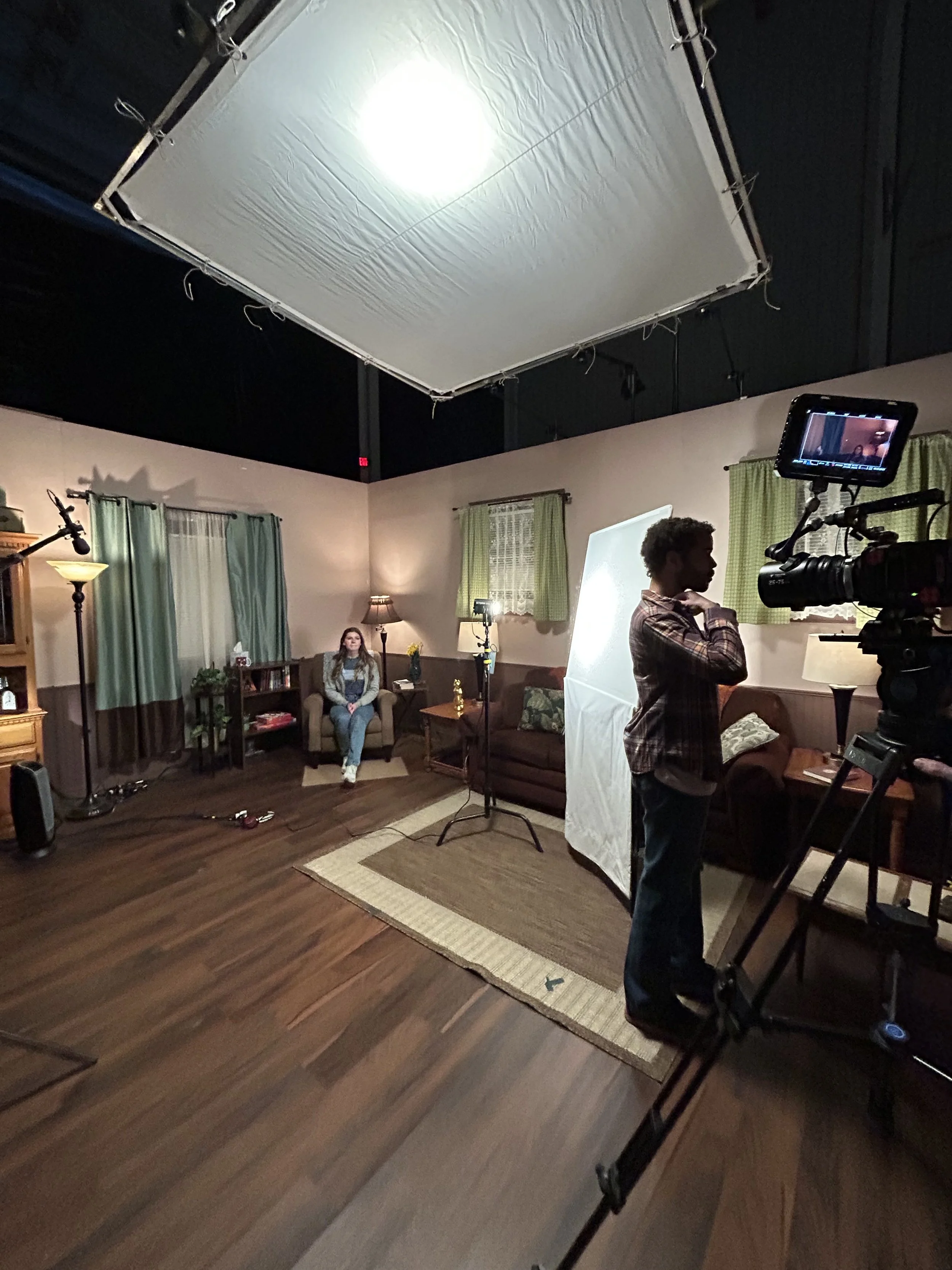 A film or video production set with a woman sitting on a couch, a crew member with a camera, lighting equipment, and a large diffuser overhead. The setting is a cozy living room with curtains, lamps, and a bookshelf.