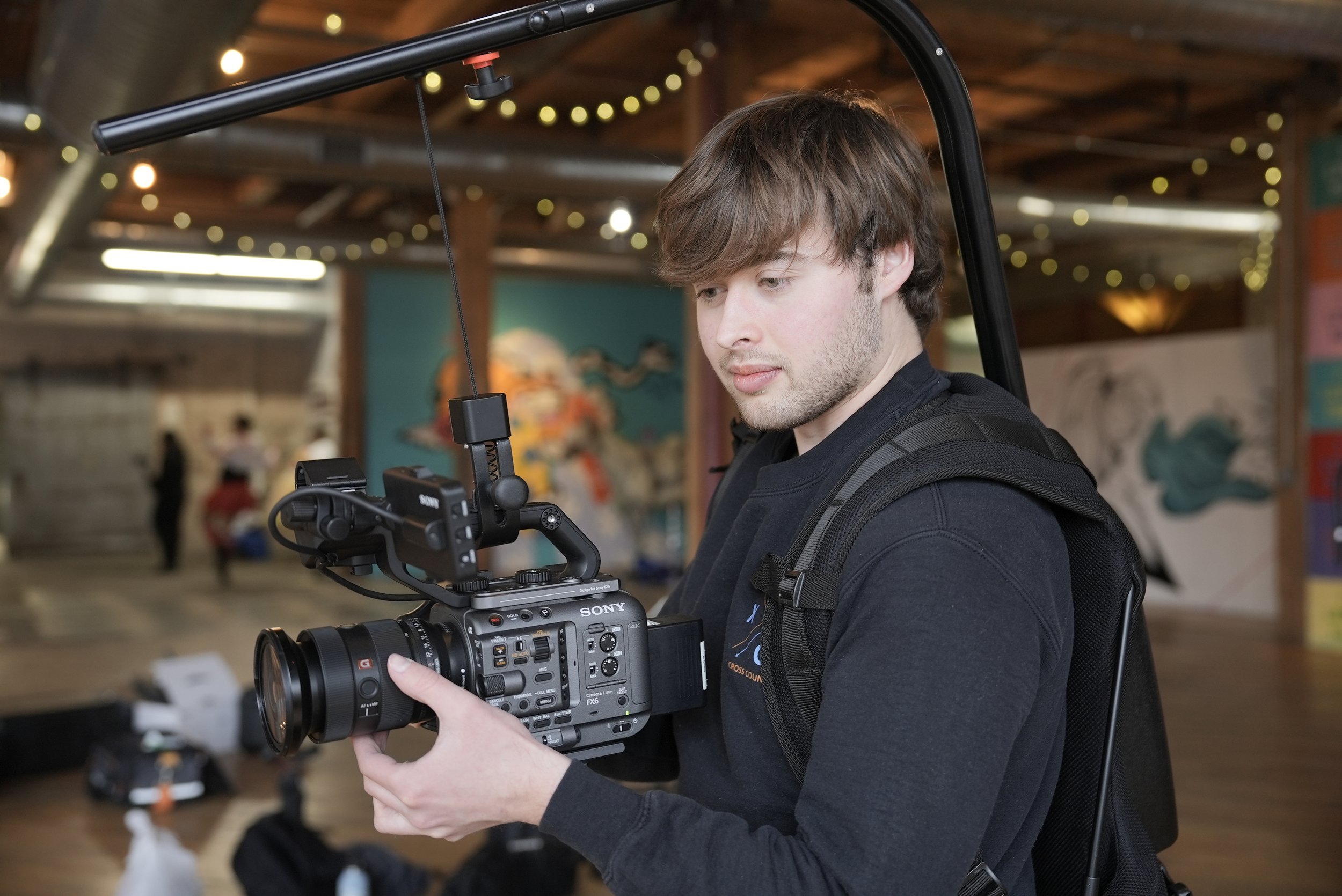 A young man operating a professional Sony video camera in an indoor setting with blurred background and string lights.