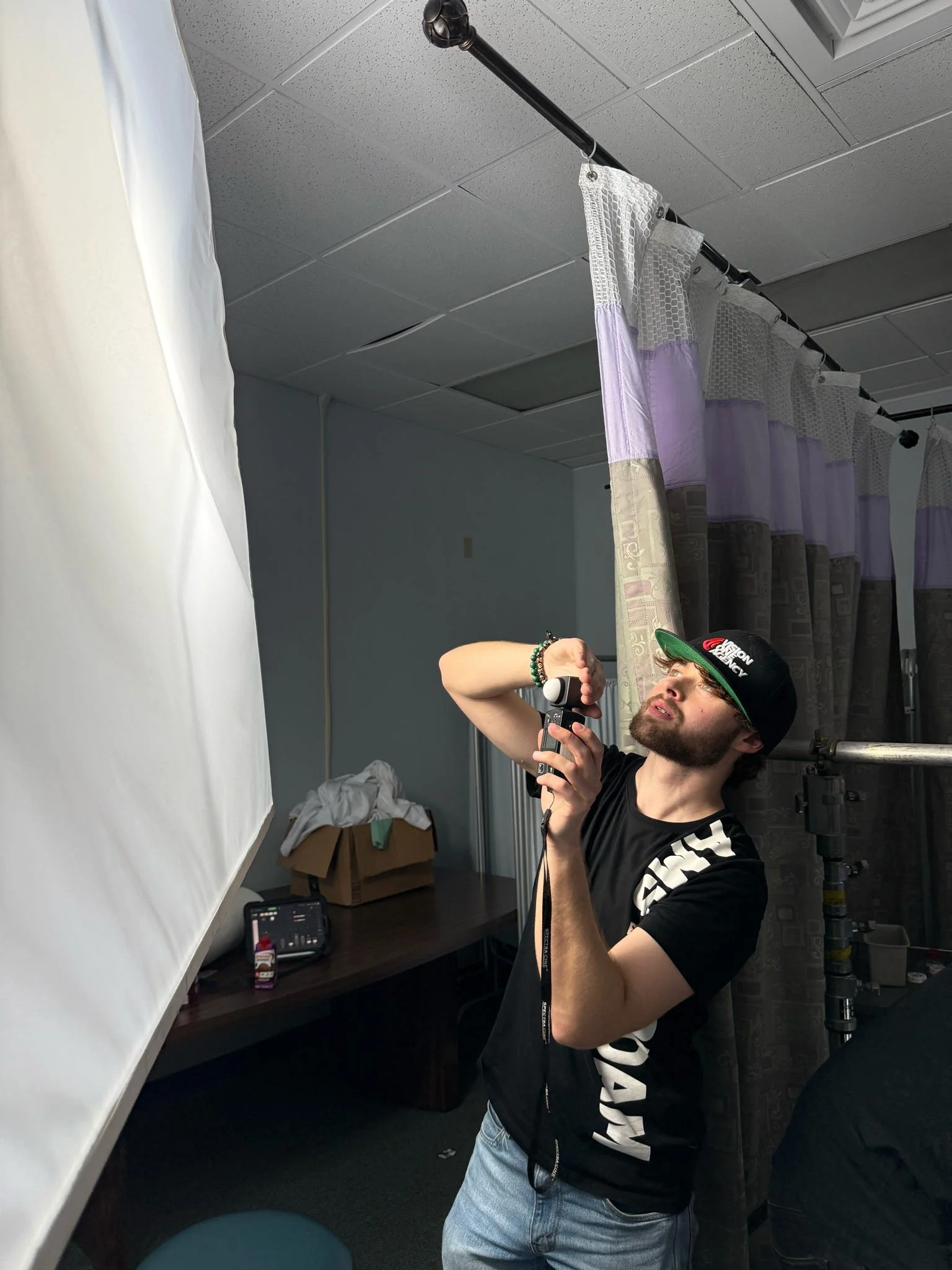 A man with a beard and wearing a black cap, black T-shirt, and jeans is adjusting a lighting reflector in a room with a white backdrop on the left. There is a table with items including a container of candy, a box, and some electronic devices. Curtains are hanging in the background.