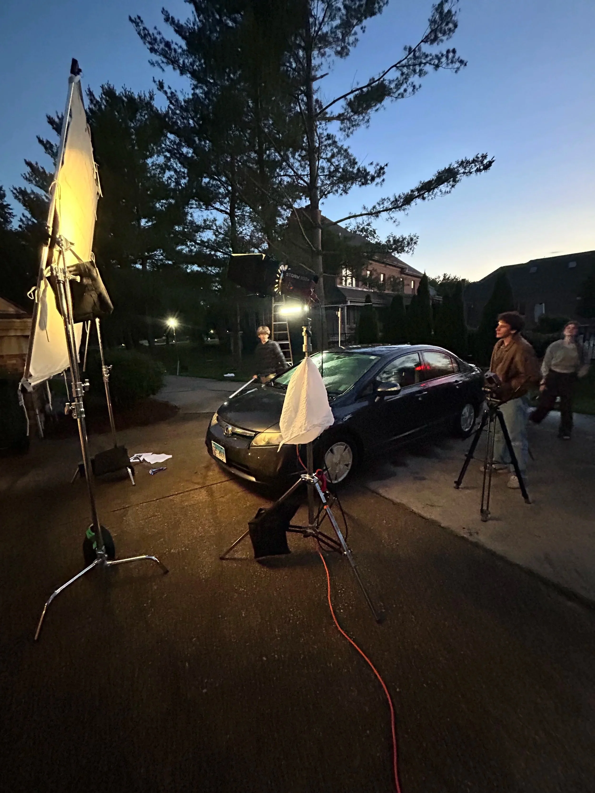 Behind the scenes of a video or film shoot at dusk, with lighting equipment, a black car, and people working, set against a neighborhood with trees and houses.