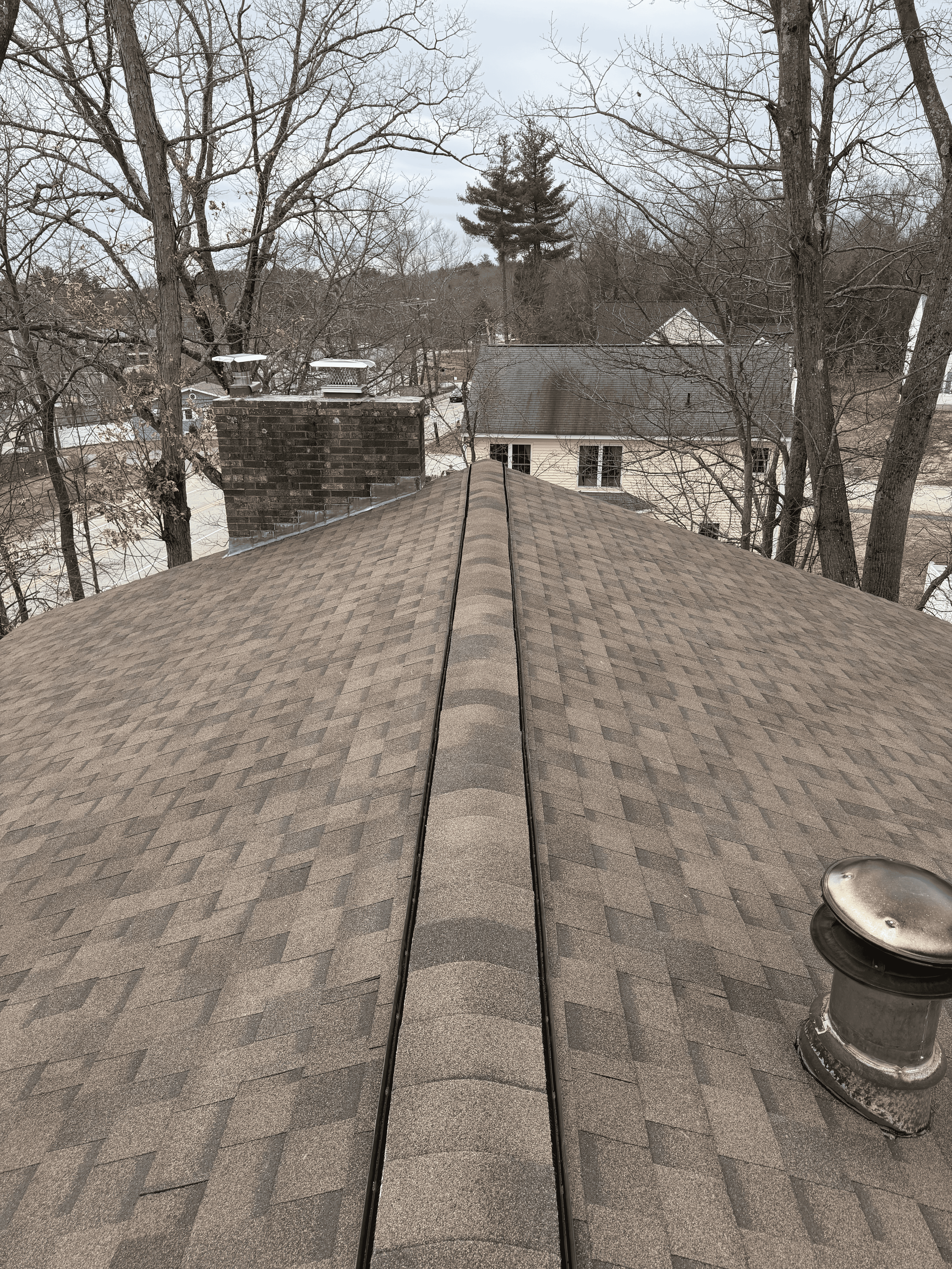 View from the roof of a house showing shingles, a chimney, and a vent pipe, with trees and neighboring houses in the background under a cloudy sky.