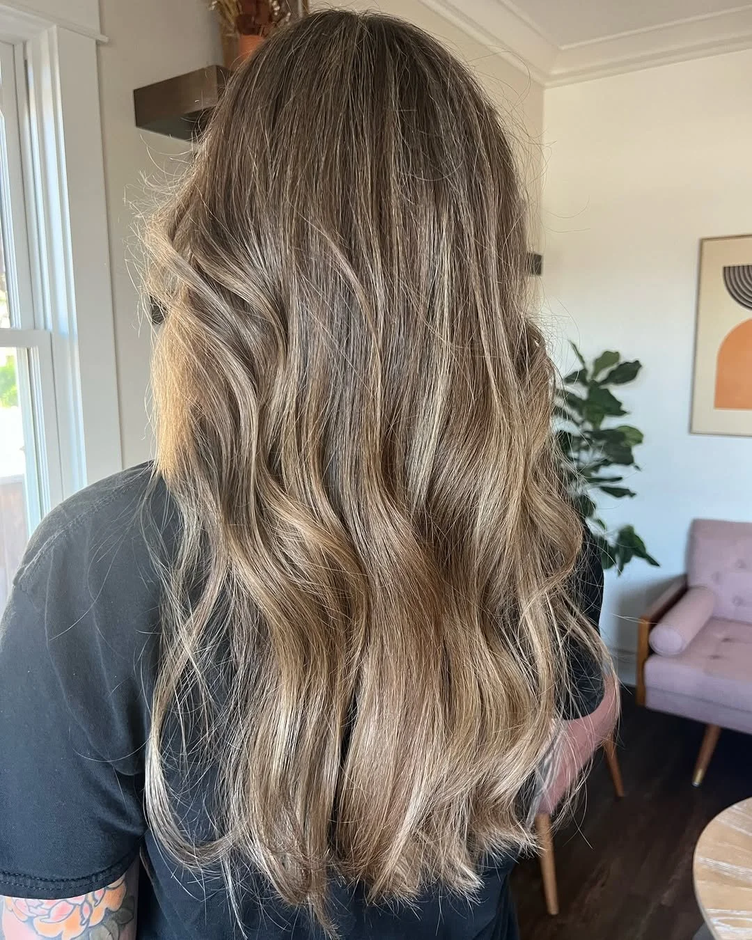 Back view of a woman with long, wavy, light brown hair in a home setting with a pink chair, a framed artwork. Hairstyle done by Caitlin Duffy Hairstylist at Bolt Collective Hair Salon in Pleasanton, CA.