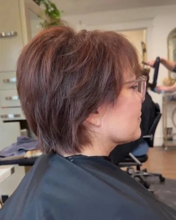 Side profile of a woman with short, layered brown hair wearing glasses, sitting in a salon chair. Hairstyle done by Caitlin Duffy Hairstylist at Bolt Collective Hair Salon in Pleasanton, CA.